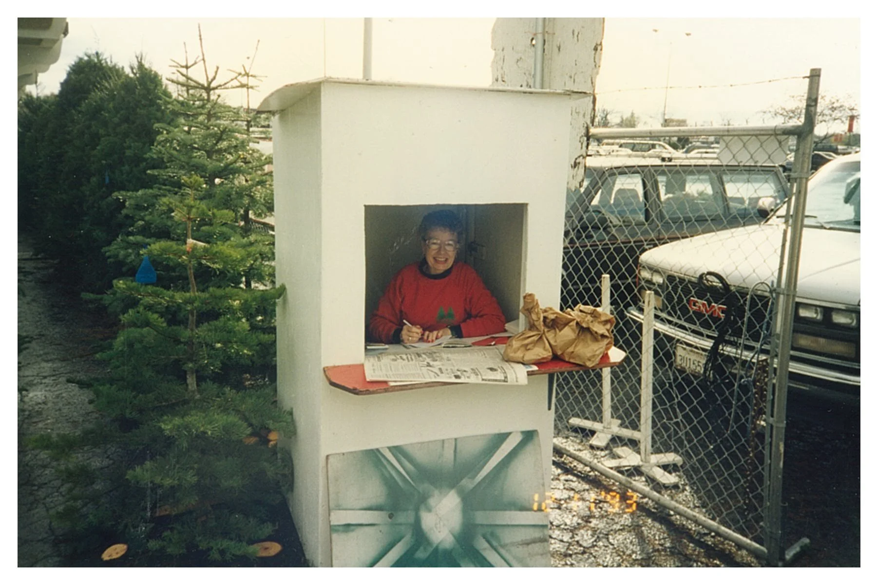 A woman sitting inside a small, white booth in a parking lot, selling fresh cut christmas trees and on a cold day. There are Christmas trees beside the booth and parked cars in the background.
