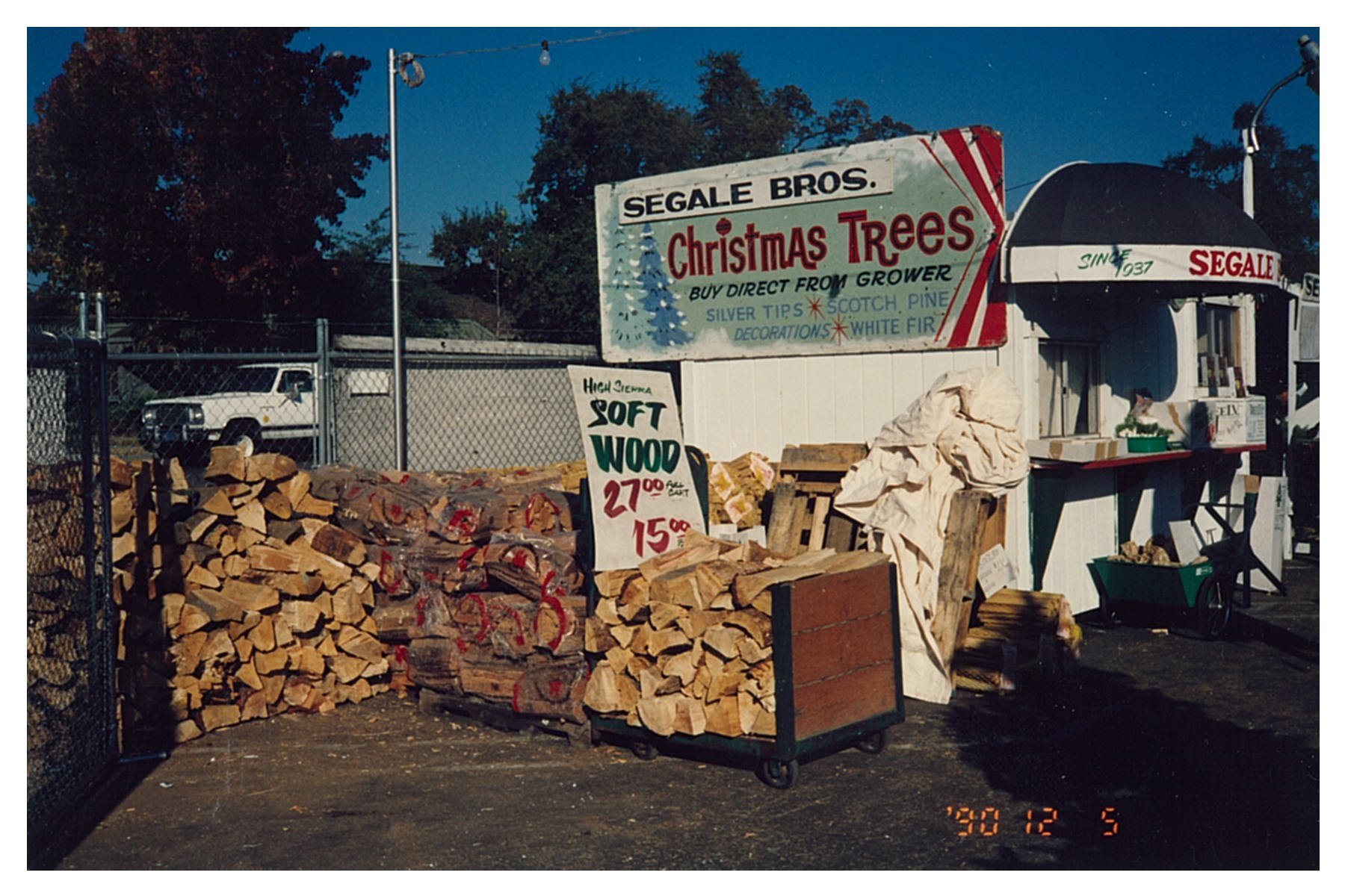 A wood stand selling firewood and Christmas trees at an outdoor market, with large stacks of cut wood and a sign with prices, under a blue sky with trees in the background.