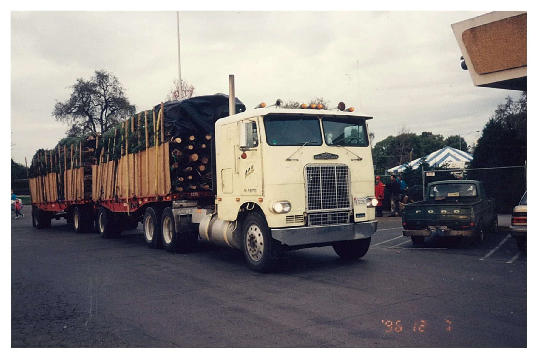 A white semi-truck carrying fresh christmas trees parked at segale christmas tree lot. Several people are near a white and blue striped tent in the background, with other cars parked around.