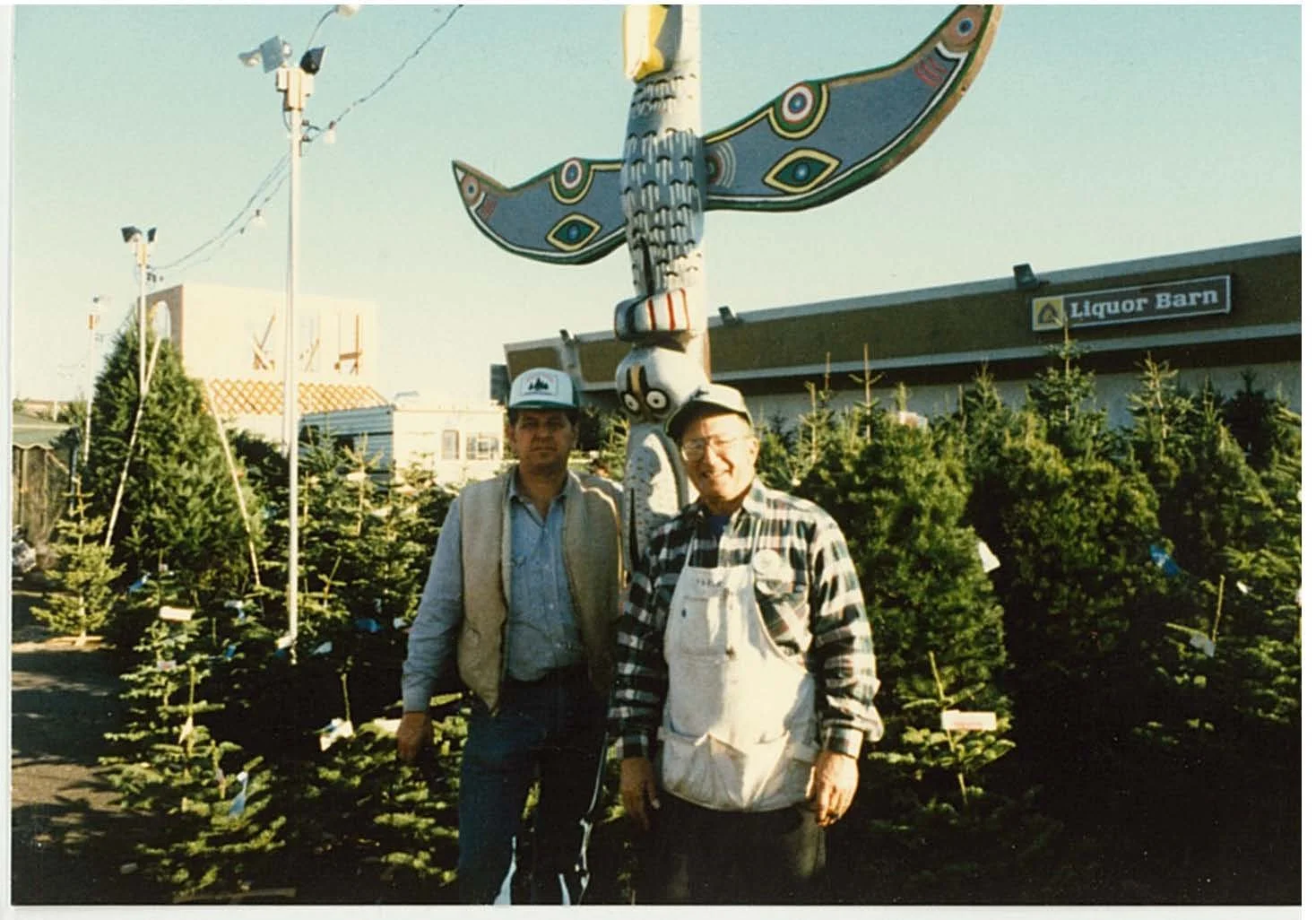 Two men standing outdoors among Christmas trees, with a decorative large butterfly sculpture mounted on a pole behind them. In the background, there is a building with a sign that reads "Liquor Barn." The scene appears to be at a Christmas tree farm or market during the daytime.