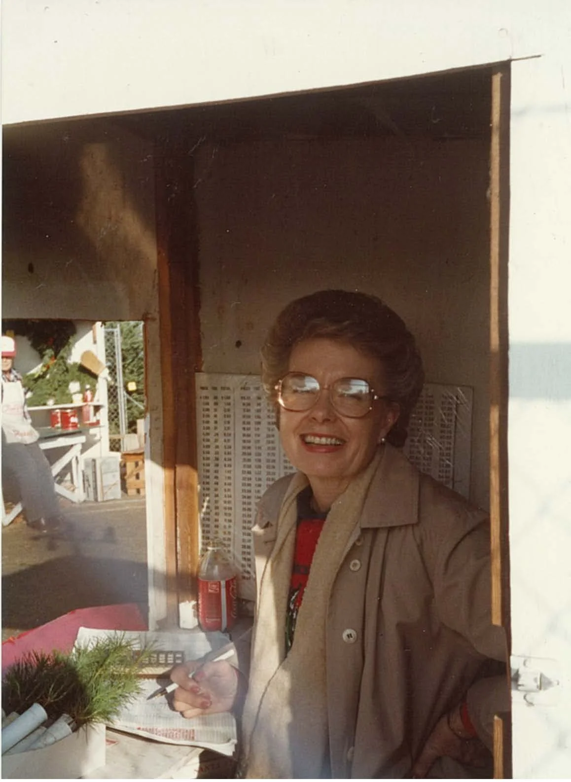 A smiling older woman with glasses and a tan jacket writing with a pen, seen through a window or opening, with holiday decorations and people in the background.