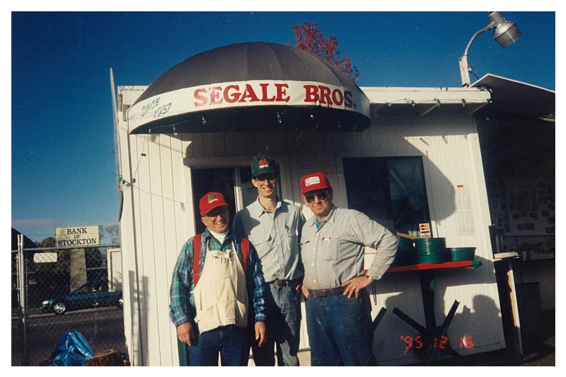 Three men standing in front of a small food stand with a sign reading 'Segale Bros' and 'Since 1937'. The men are smiling and wearing hats, with one wearing an apron. The stand is outdoors with a fence and a sign for 'Bank of Stockton' visible in the background. The date on the photo is December 6, 1995.