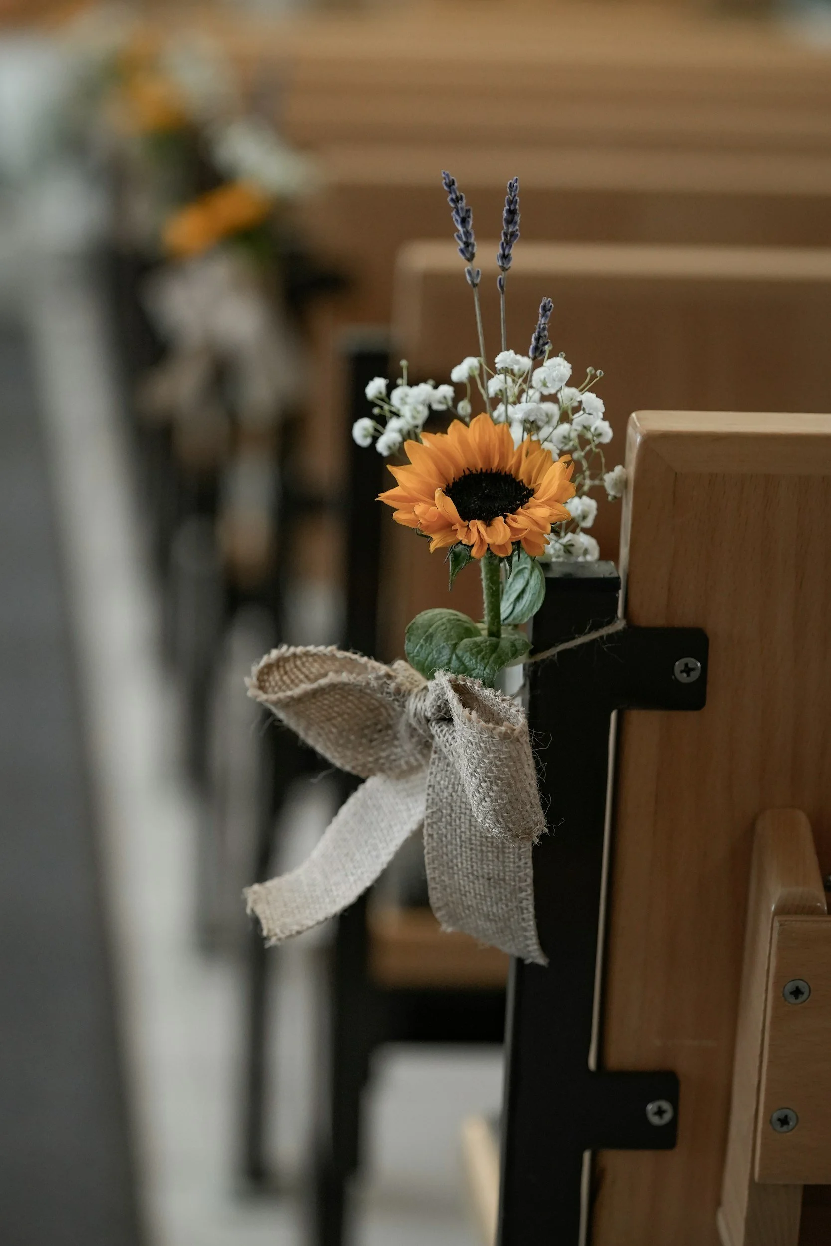 The image shows a small flower arrangement attached to the side of a wooden piano. The arrangement includes a sunflower, white baby's breath, and purple lavender, with a burlap ribbon tied around the base.
