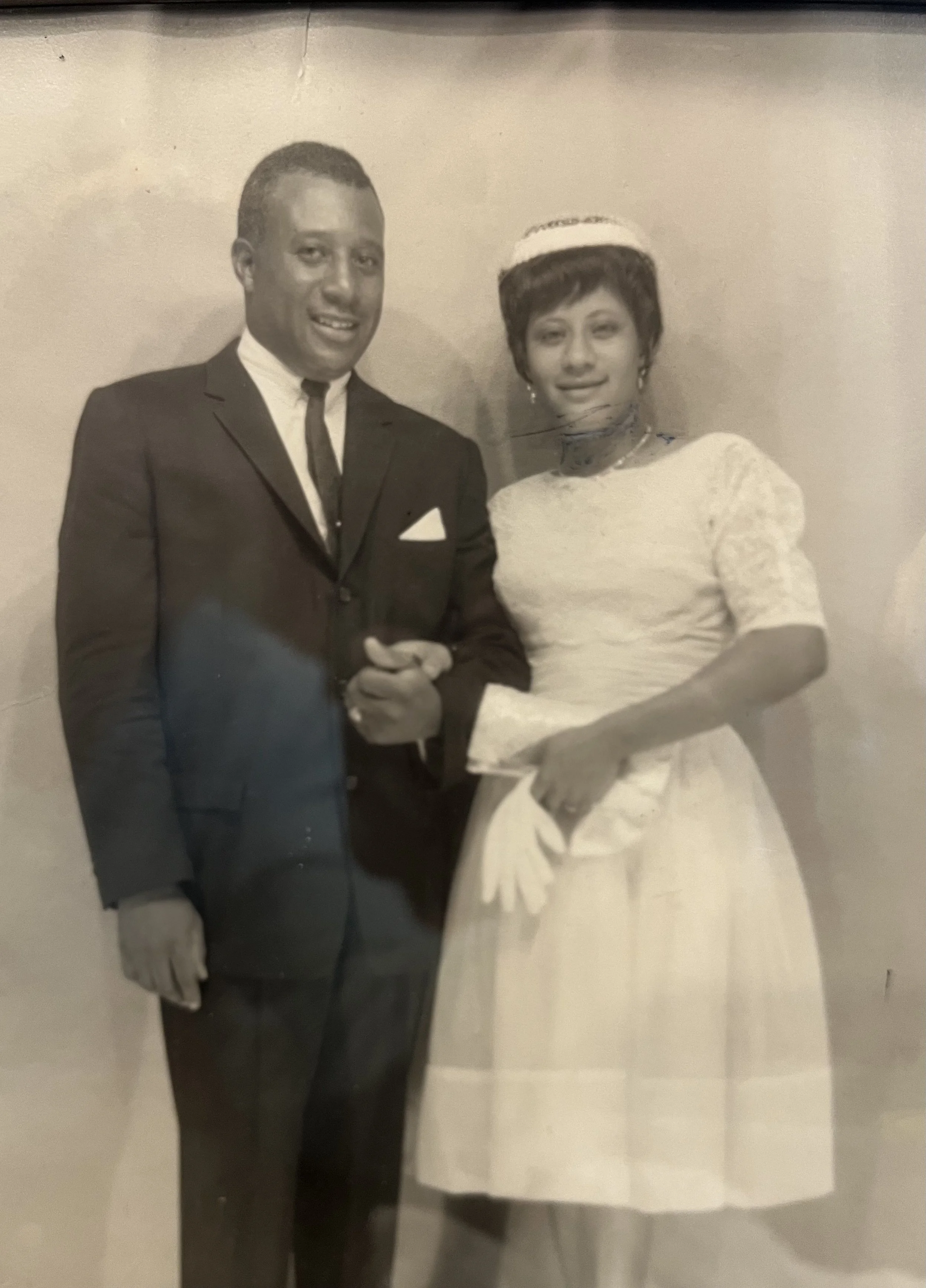 A black and white photo of a man in a suit and a woman in a dress holding hands, standing together, with the woman wearing a head covering and earrings.