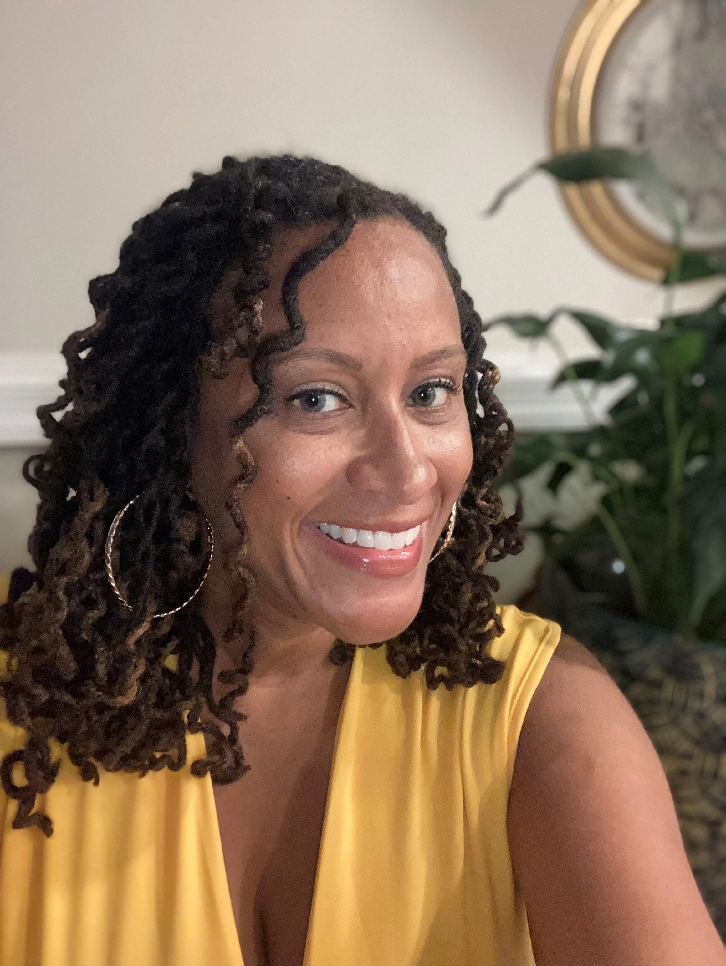 Close-up of a woman with dark curly hair, smiling, wearing a yellow sleeveless top, in front of a white wall and a green plant.