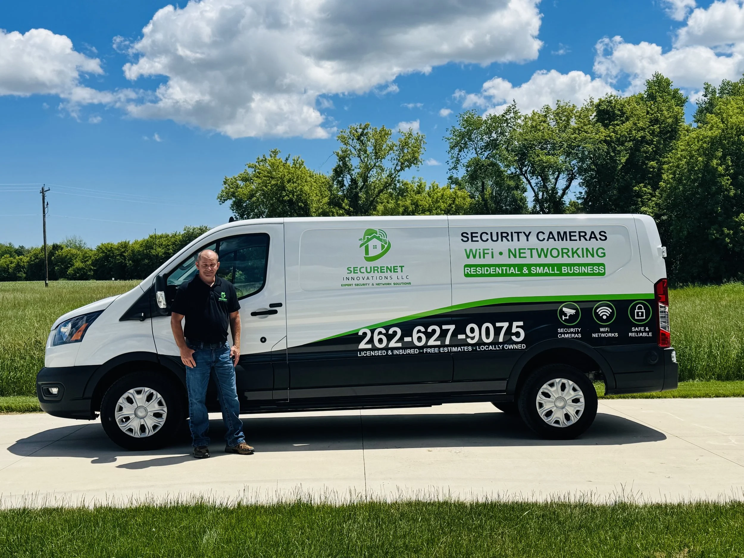 A man standing next to a white van with advertising for security cameras, WiFi, and networking services parked on a driveway in front of a grassy area with trees and a blue sky with clouds.