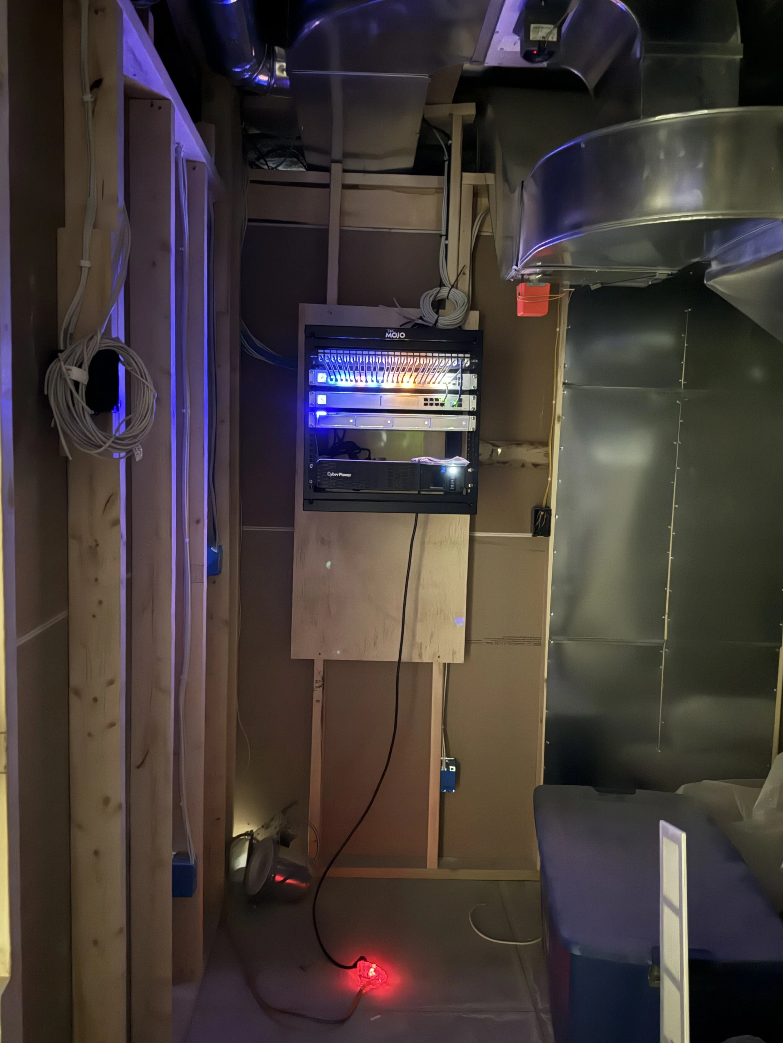 The image shows a partially constructed bar or countertop with wooden framing, electrical wiring, and a server rack. There are metallic surfaces to the right, and the scene is dimly lit with some colored LED lights.