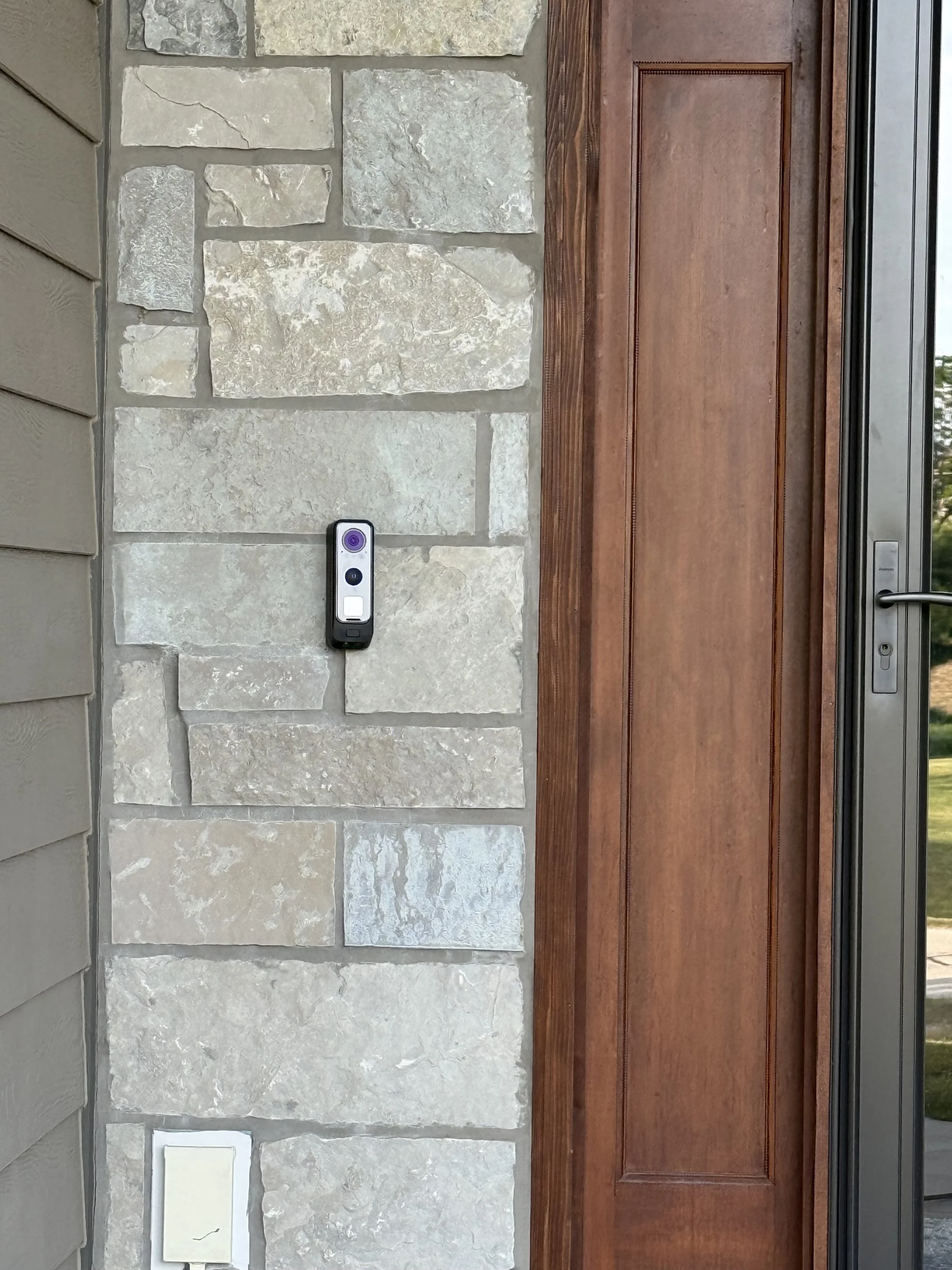 Close-up of a front door with a stone wall and wooden frame, with a doorbell camera below a light. The door has a metal handle and a visible lock.