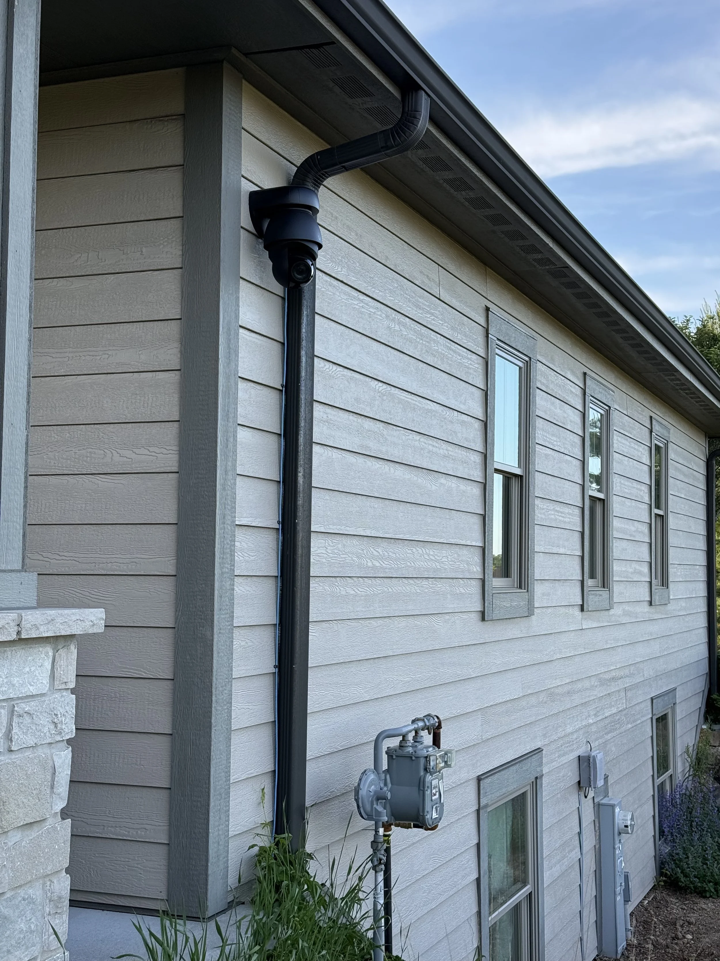 View of the side of a house with beige horizontal siding, three windows, and an overhanging roof. A security camera and gas meter are attached to the wall, with a black downspout running from the roof to the ground.