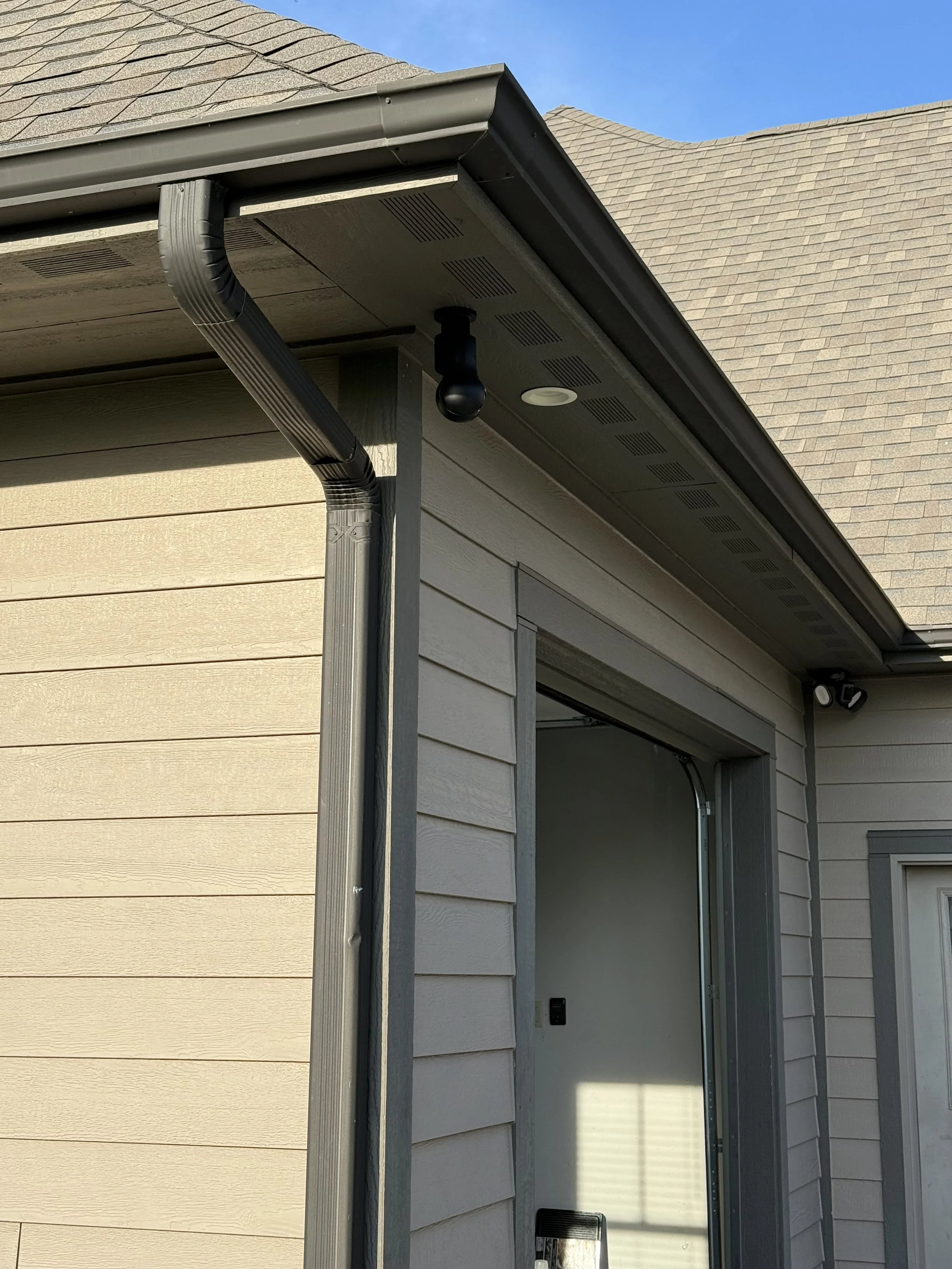 Close-up of a house exterior showing beige horizontal siding, a partial view of a garage with an open door, a rain gutter, and a security camera.