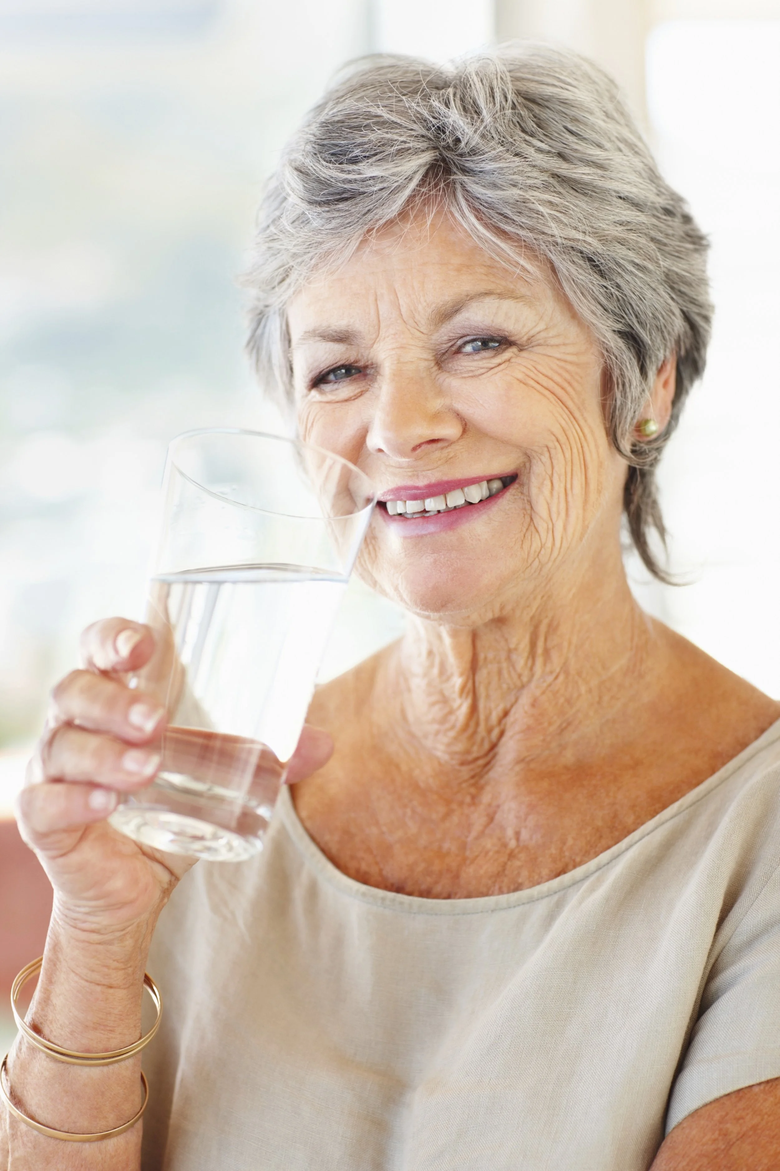 Close-up of an elderly woman with gray hair smiling and holding a glass of water, wearing a beige top, gold earrings, and gold bangles.
