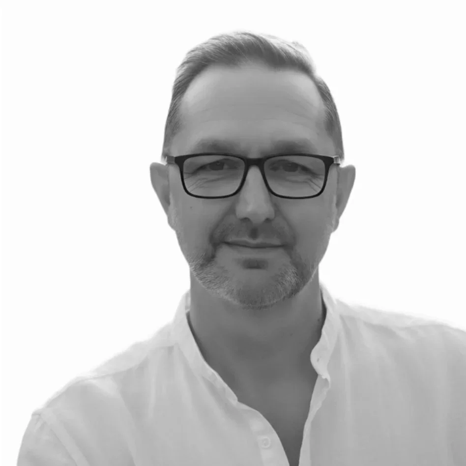Black and white portrait of a man wearing glasses and a collared shirt, smiling gently, against a plain white background.