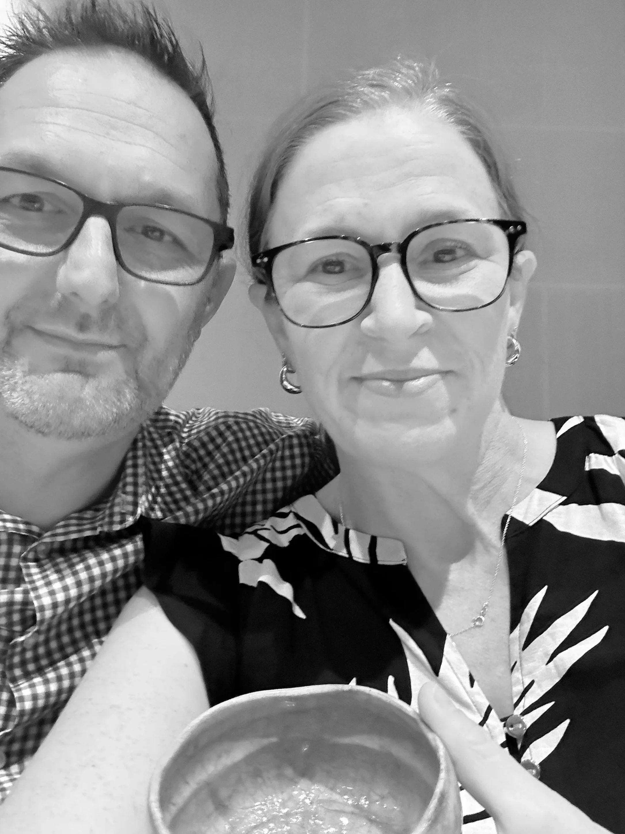 A black-and-white photo of a man and a woman taking a selfie together, both wearing glasses. The man's face is partially visible on the left side, with spiked hair and a checked shirt. The woman is on the right, smiling slightly, wearing earrings and a patterned top. They are positioned close to each other, and a mug is visible at the bottom of the image.