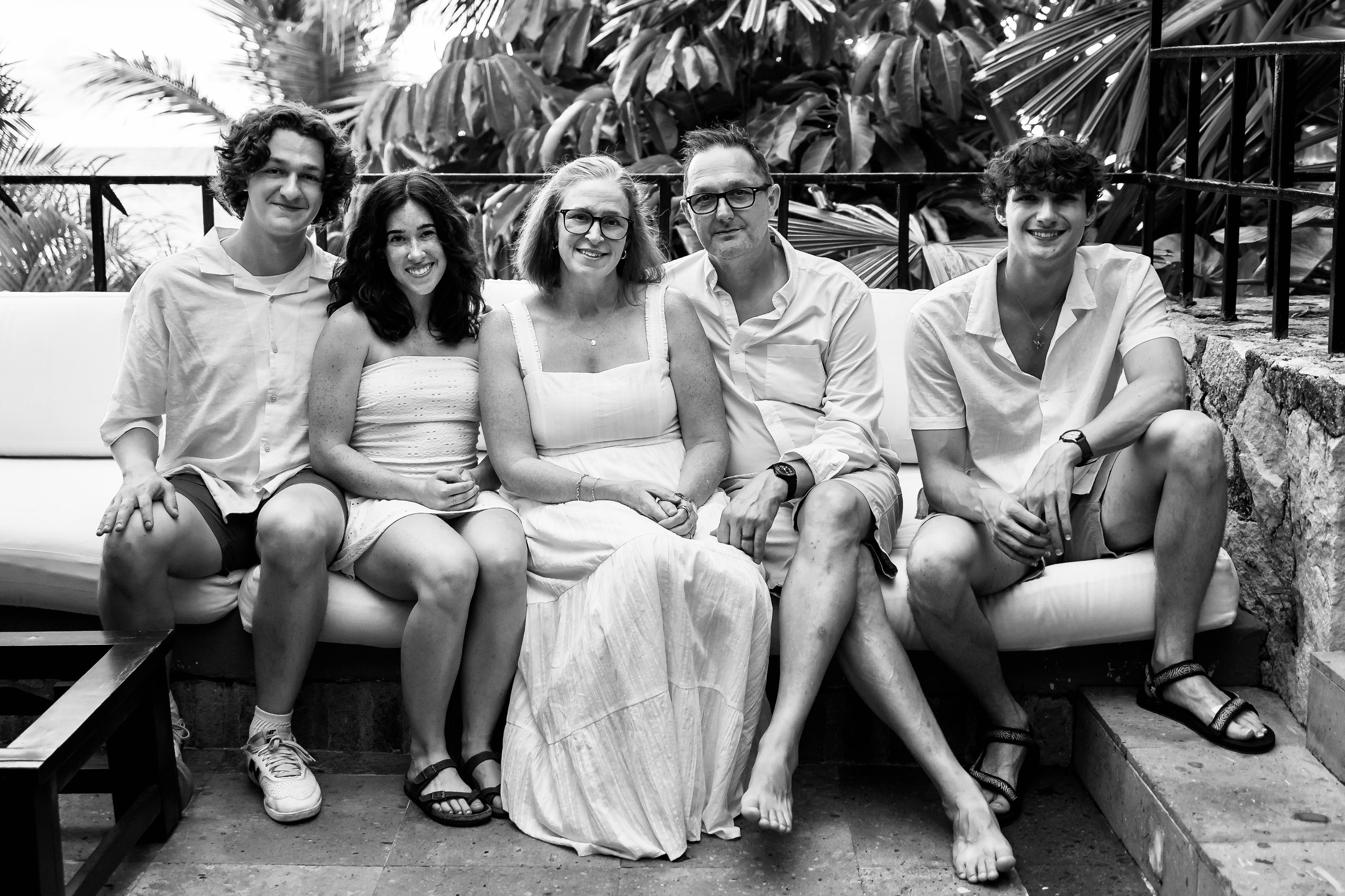 Black and white photo of five people sitting on a couch in front of large tropical leaves. Four are smiling at the camera, one has a neutral expression. They are dressed in casual summer clothing.