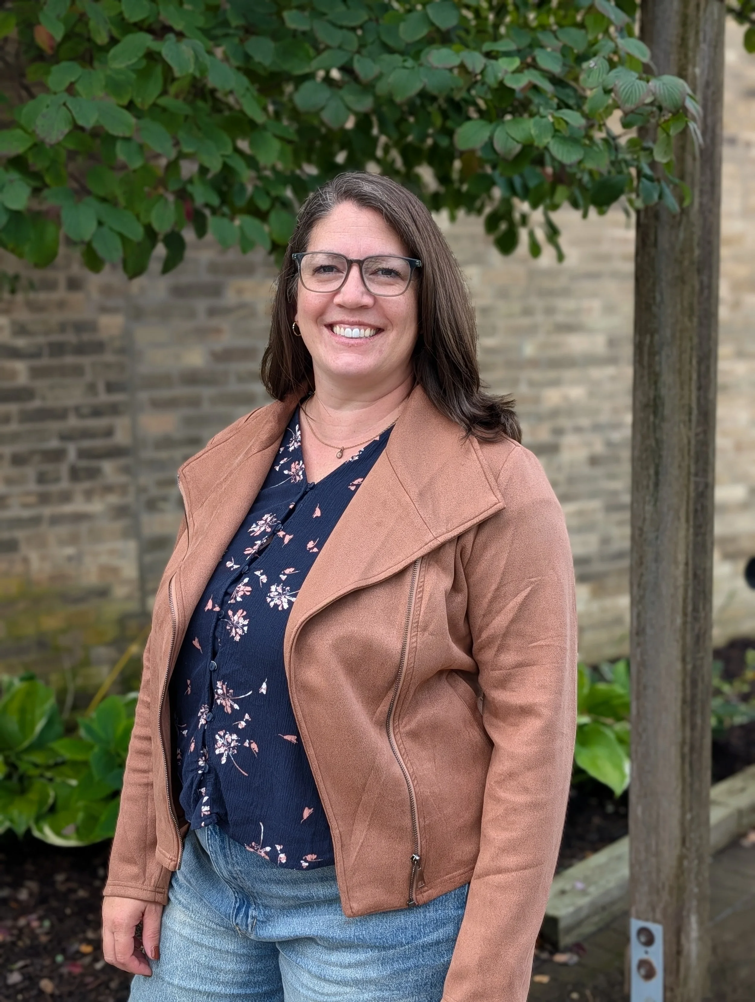 A woman with shoulder-length brown hair and glasses standing outdoors in front of a tree with green leaves and a brick wall, smiling at the camera.