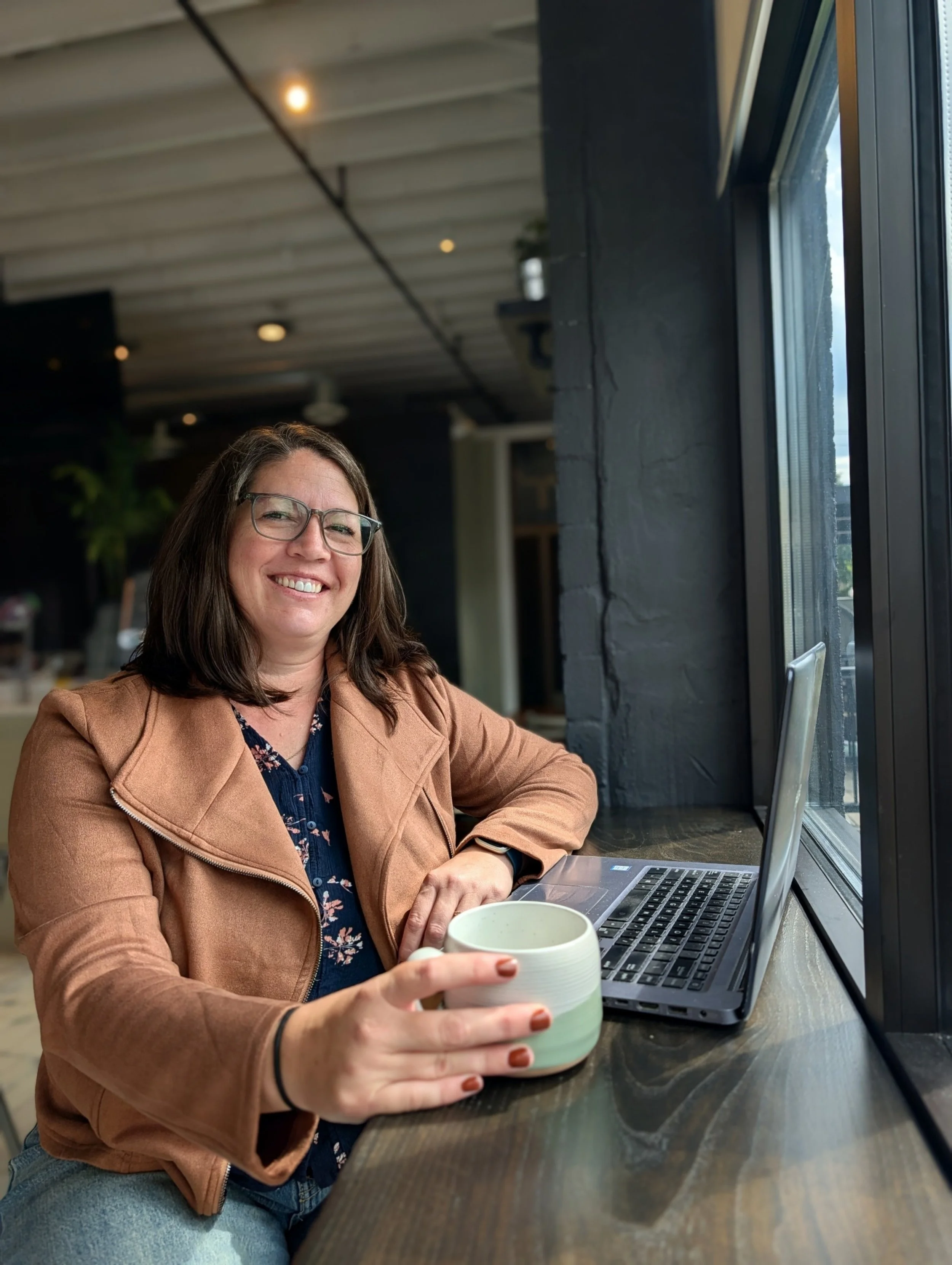 A woman with glasses smiling while sitting at a wooden table in a café, holding a white mug, with a laptop open in front of her near a large window.