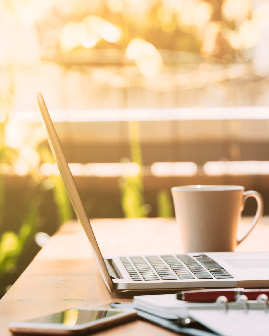 A workspace with a laptop, a coffee mug, a smartphone, and a notebook on a wooden table, with sunlight and greenery in the background.