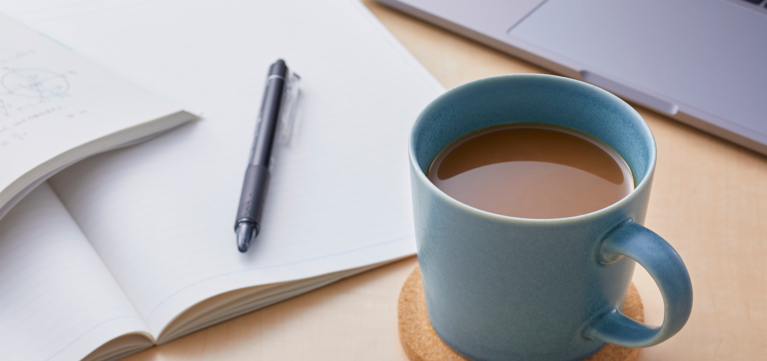 A blue ceramic mug filled with coffee sitting on a cork coaster on a light wooden desk. Nearby, an open notebook with a black pen resting on it, and a closed laptop in the background.