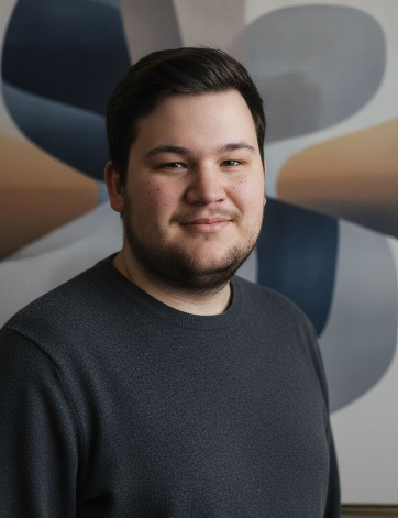 A young man with short dark hair and a beard, wearing a dark gray shirt, standing in front of a colorful abstract background.