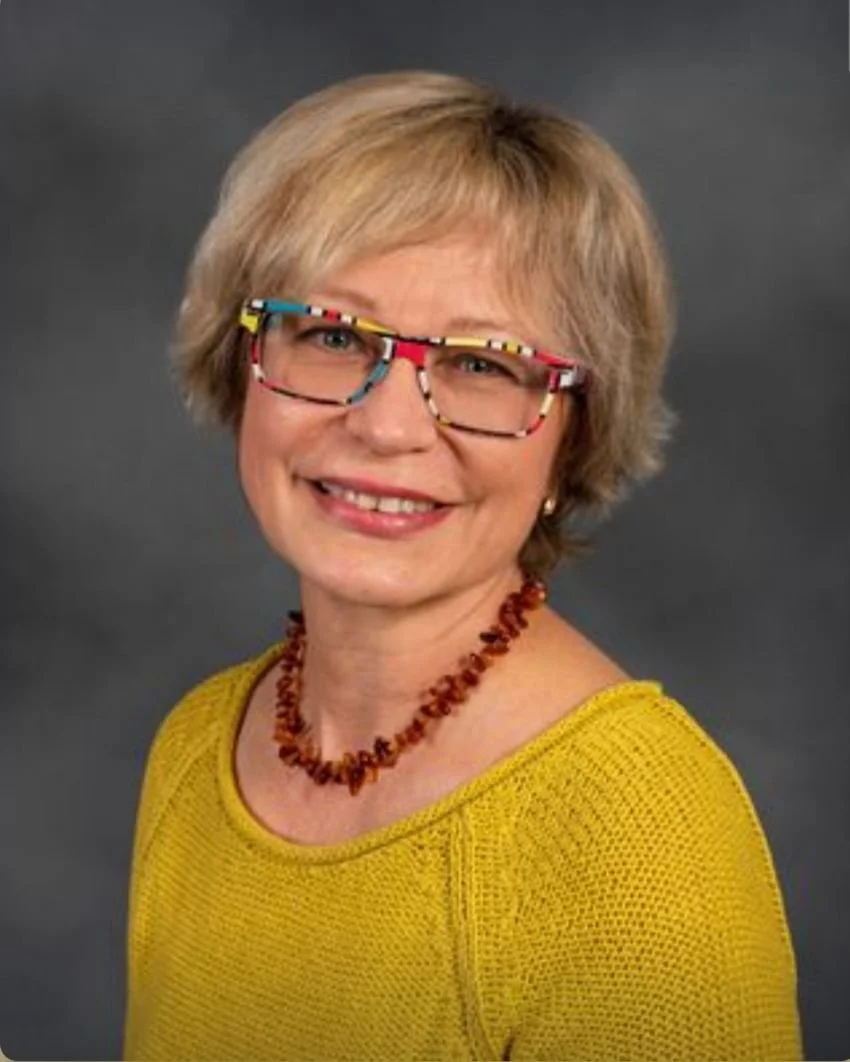 A woman with short blonde hair, wearing colorful glasses, a yellow sweater, and a reddish-brown beaded necklace, smiling at the camera against a gray background.