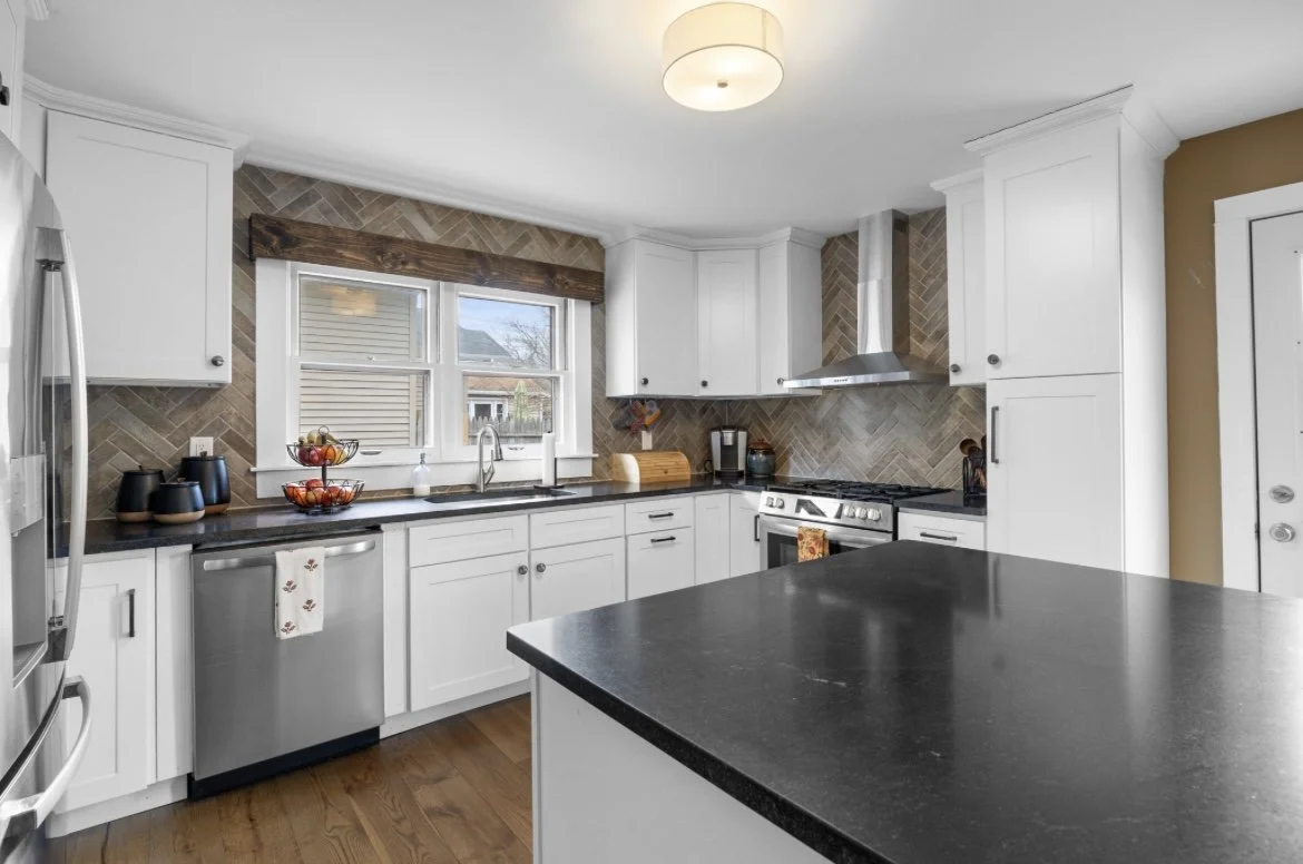A modern kitchen with white cabinets, a black countertop island, stainless steel appliances, a tile backsplash, and a window above the sink.