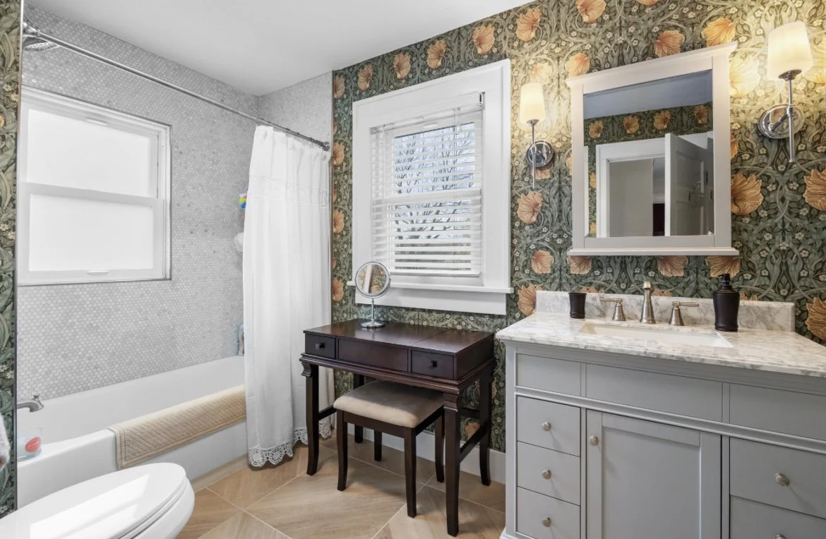 A vintage-style bathroom with floral wallpaper, a white vanity with marble countertop, a mirror, wall sconces, a window with blinds, a small desk with a stool, and a bathtub with a shower curtain.