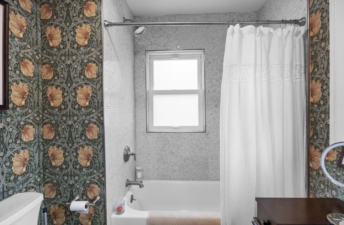 Bathroom with floral patterned wallpaper, a bathtub with a showerhead, a small window, a white shower curtain, and a brown wooden cabinet.