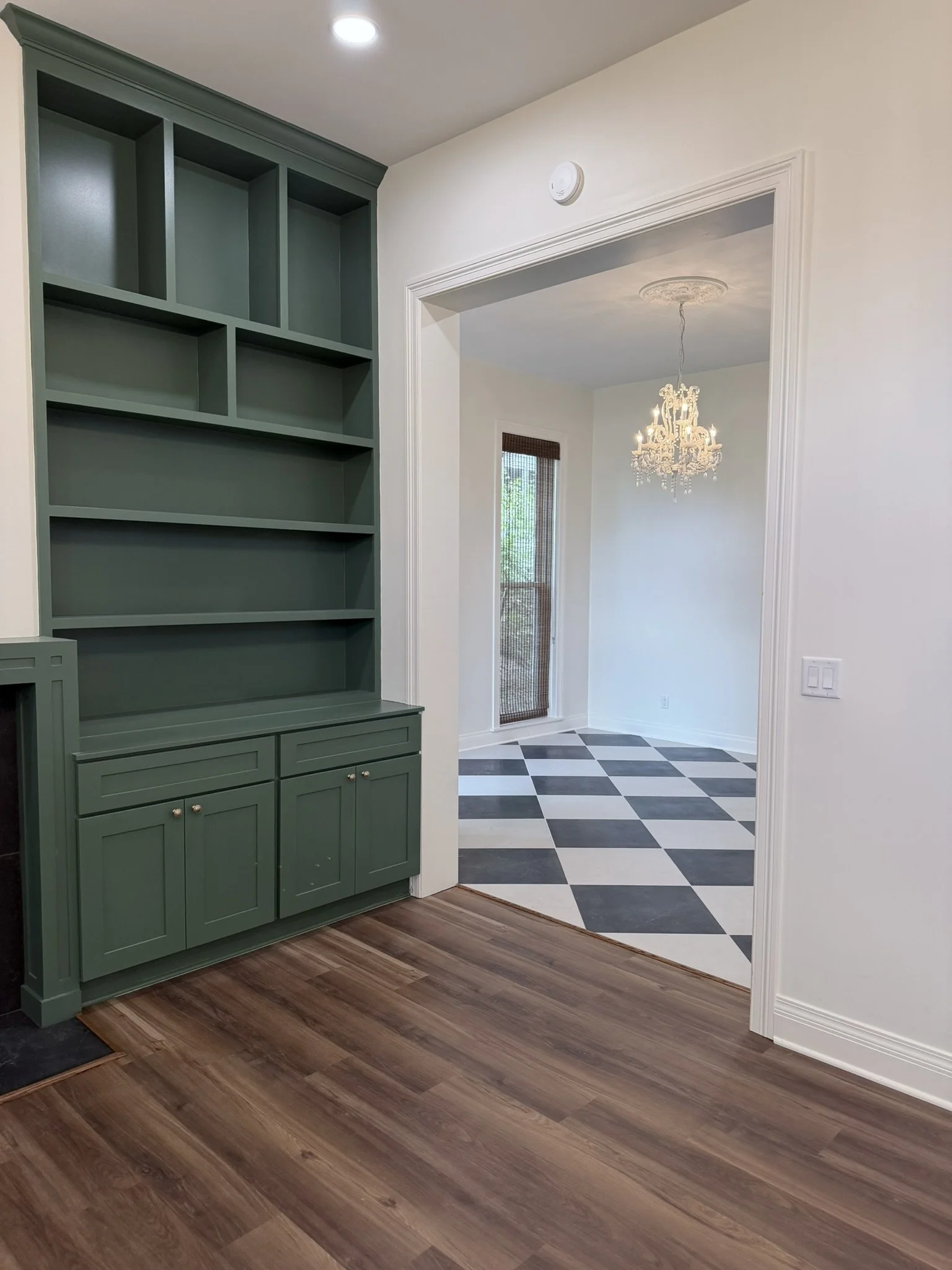 View of an interior room with a green built-in bookshelf and a doorway leading to a space with a chandelier and checkered black and white floor tiles.