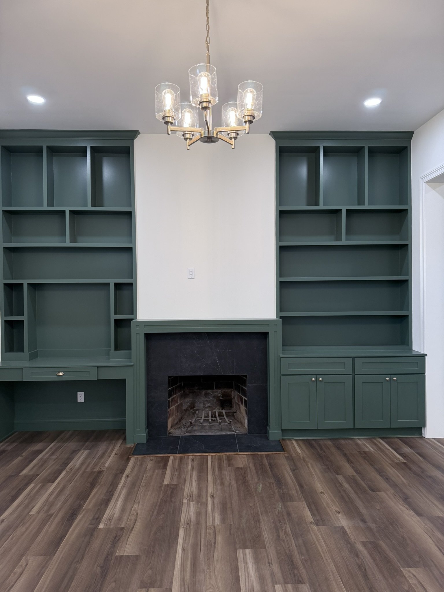 Living room with a fireplace, built-in bookshelves painted green on either side, a chandelier hanging from the ceiling, and wood flooring.