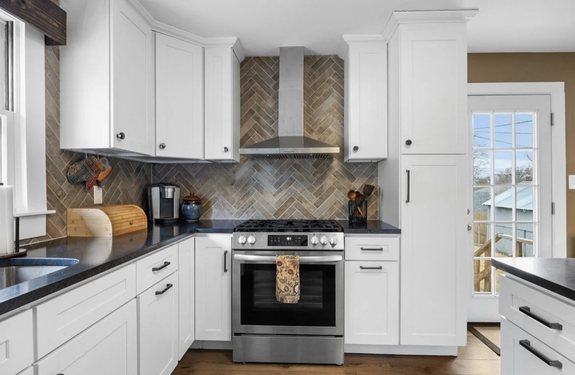 Modern kitchen with white cabinets, black countertops, a stainless steel stove, and a herringbone tile backsplash, with a glass door leading outside.