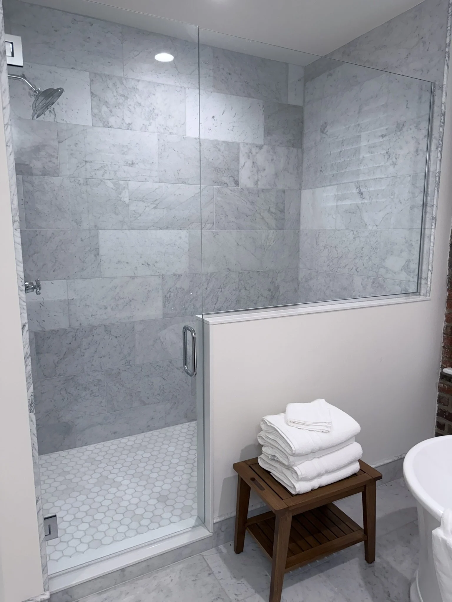 A bathroom shower with a glass door, marble tiles, and a showerhead. There are white towels neatly folded on a wooden bench and a small section of a white toilet is visible.