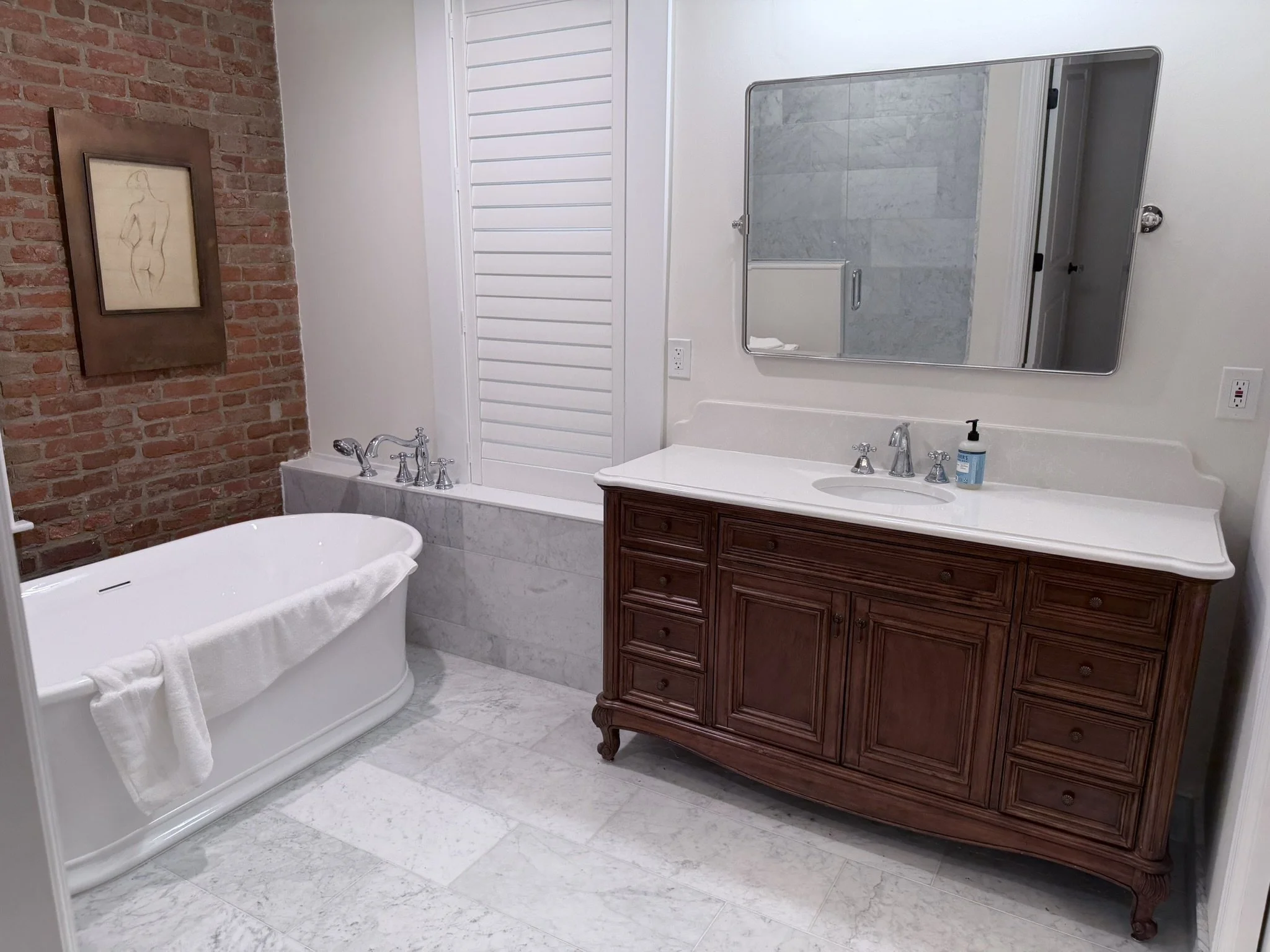 A bathroom with a white bathtub, a wooden vanity with a mirror, and a brick wall with a framed sketch of a nude woman. The floor and part of the wall are covered in white marble tiles.