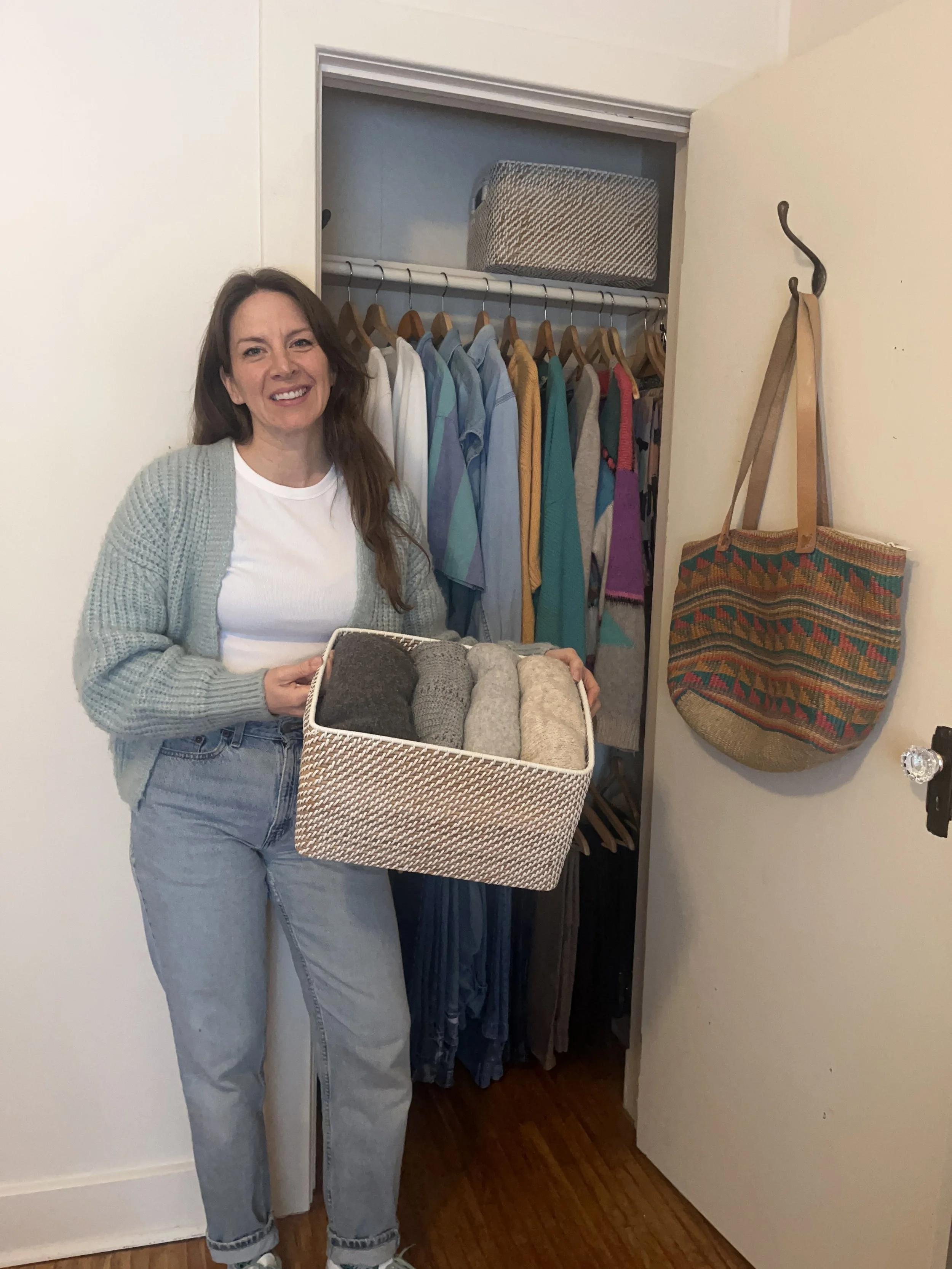 A woman standing near an open closet holding a basket of rolled textiles, with clothes hanging inside the closet and decorative bags on the wall.