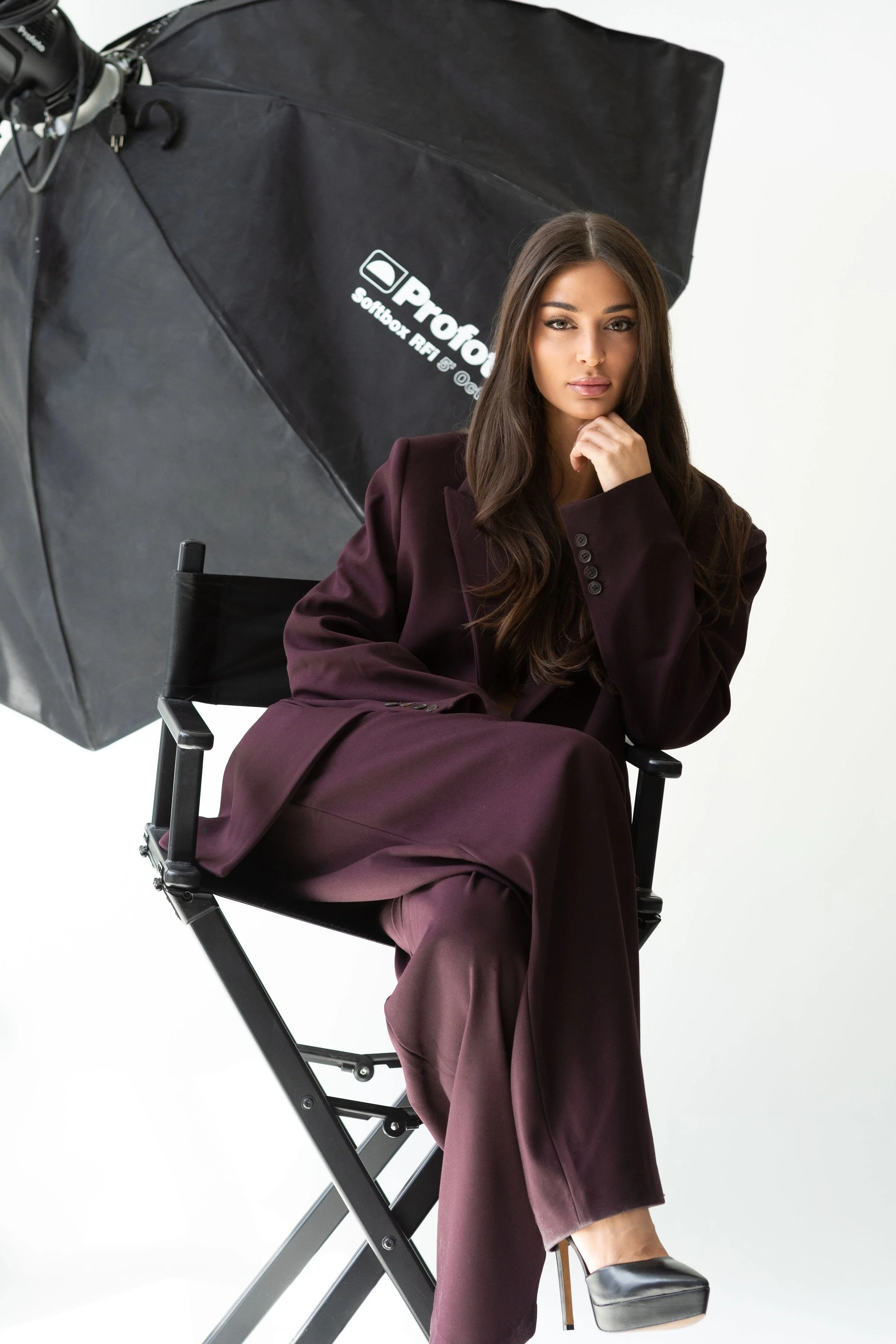 A woman with long dark hair sitting on a director's chair in a black suit and high heels, posing for a photo in a studio with a large professional softbox.