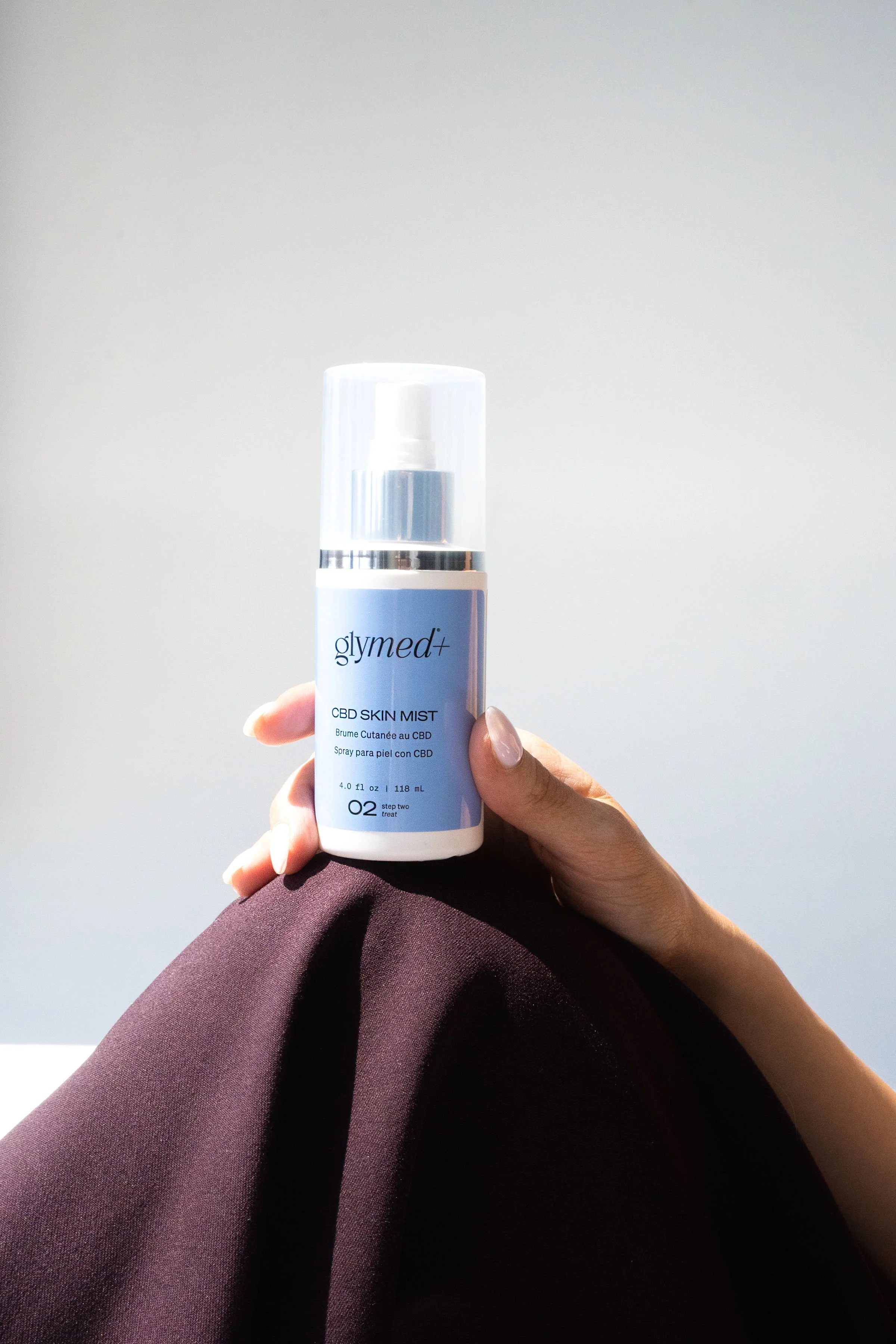 A person holding a white bottle of Glymed CBD Skin Mist with a spray nozzle, with a neutral background and wearing dark clothing.