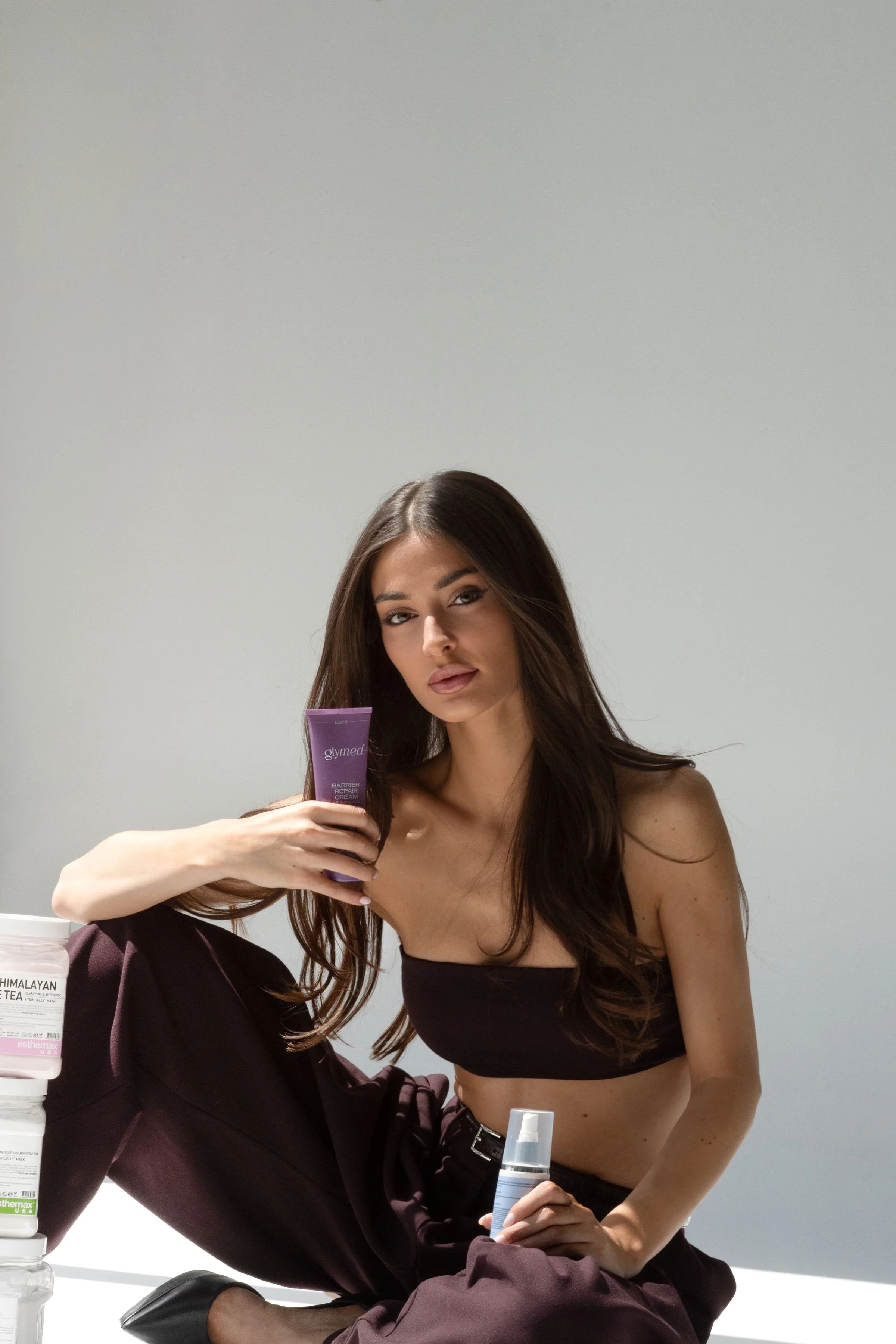 A woman with long brown hair, wearing a black crop top, is sitting and holding a purple skincare product. She is seated next to another person in dark pants, whose face is not visible, holding a small white spray bottle. The background is a plain white wall.