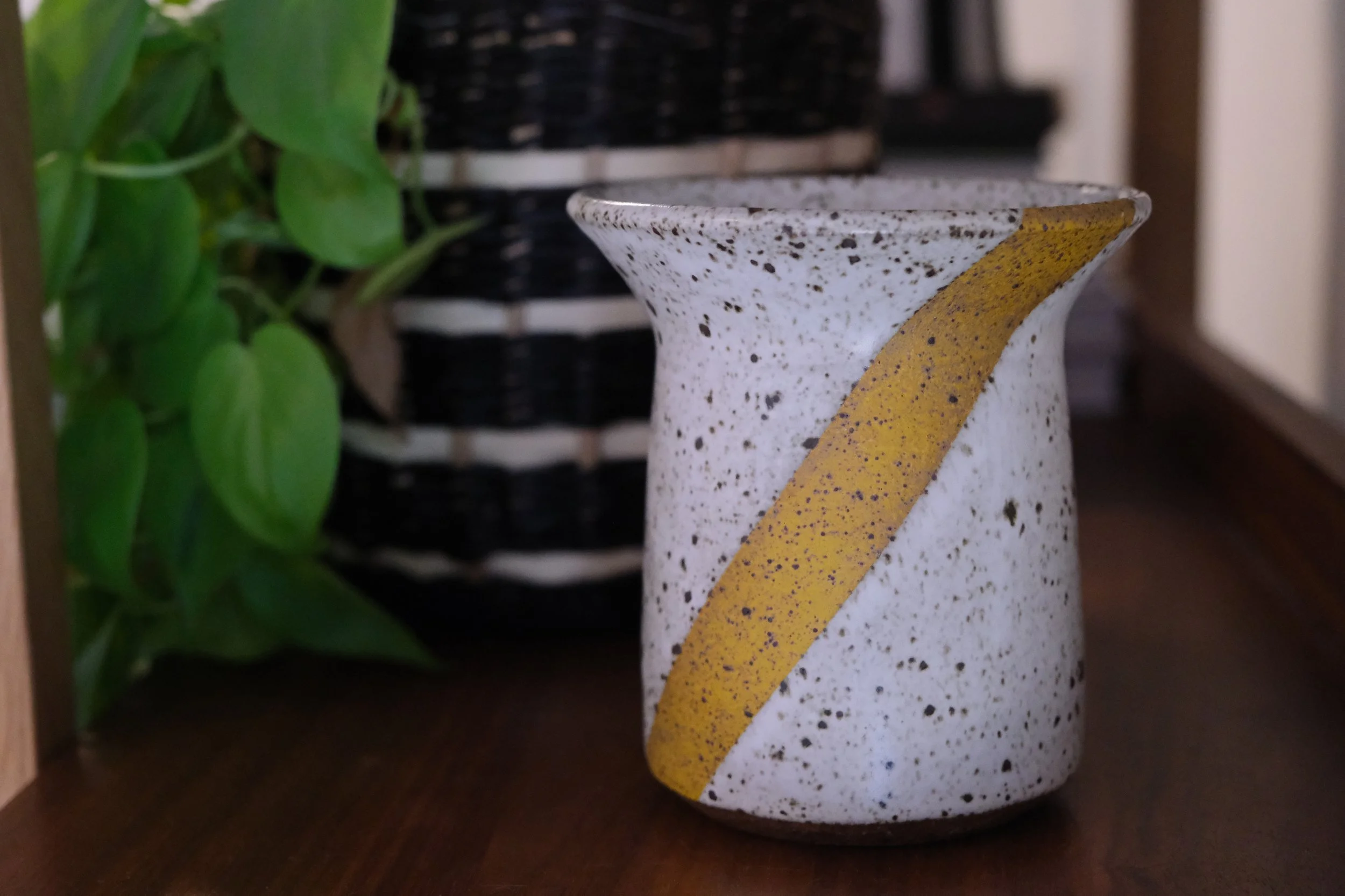 Close-up of a speckled ceramic vase with a yellow stripe, positioned on a wooden surface, with green leaves nearby.