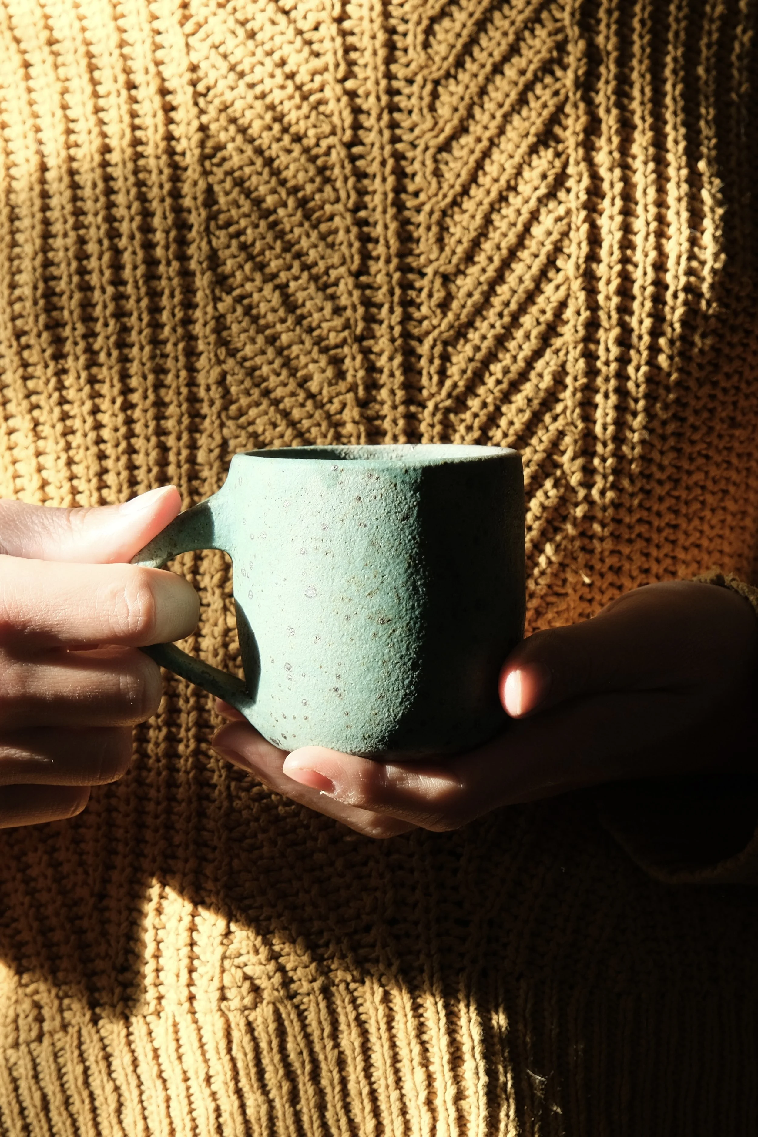 Two hands holding a light turquoise ceramic mug against a yellow textured knit background.