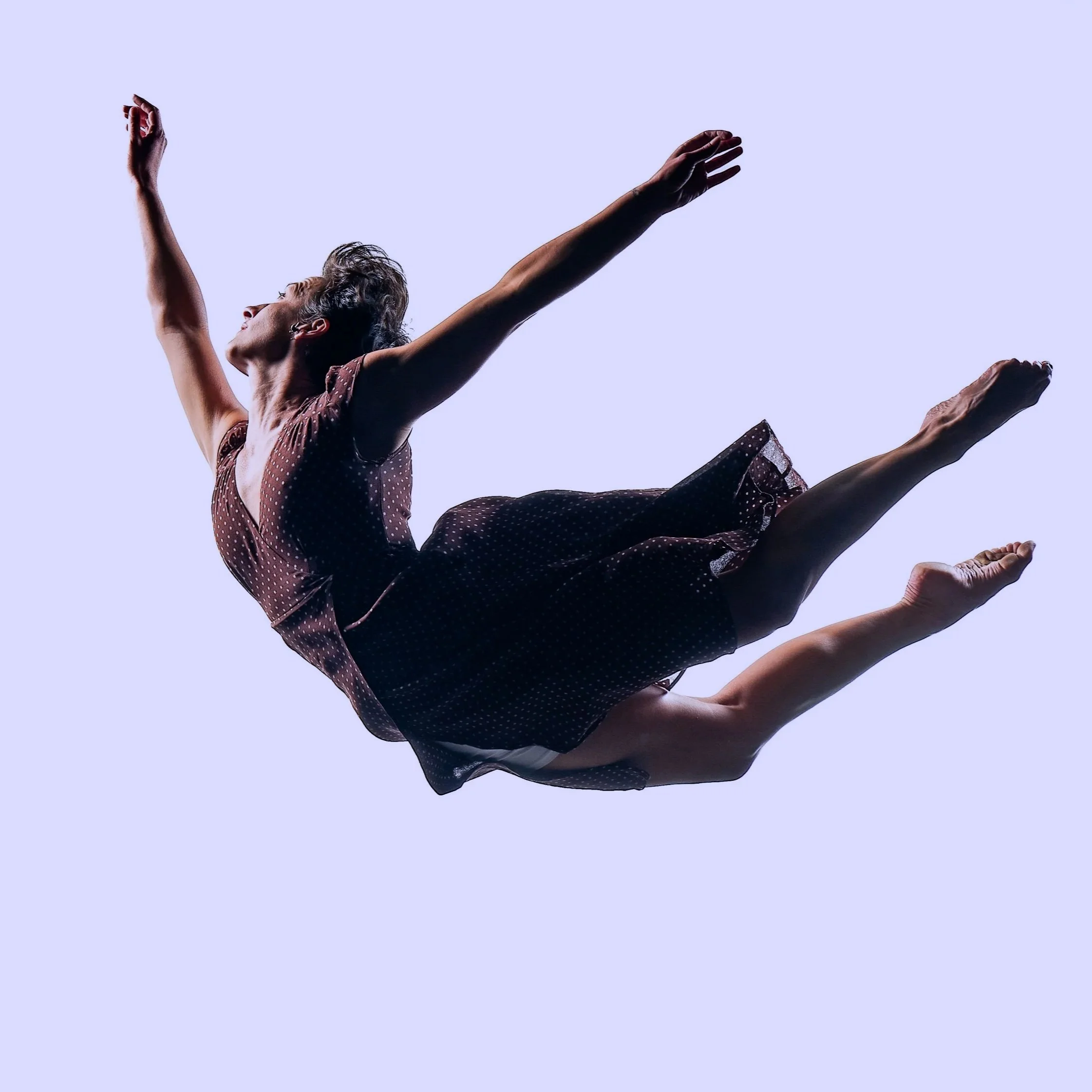A dance performer in mid-air with arms outstretched, wearing a sleeveless outfit, set against a light purple background.