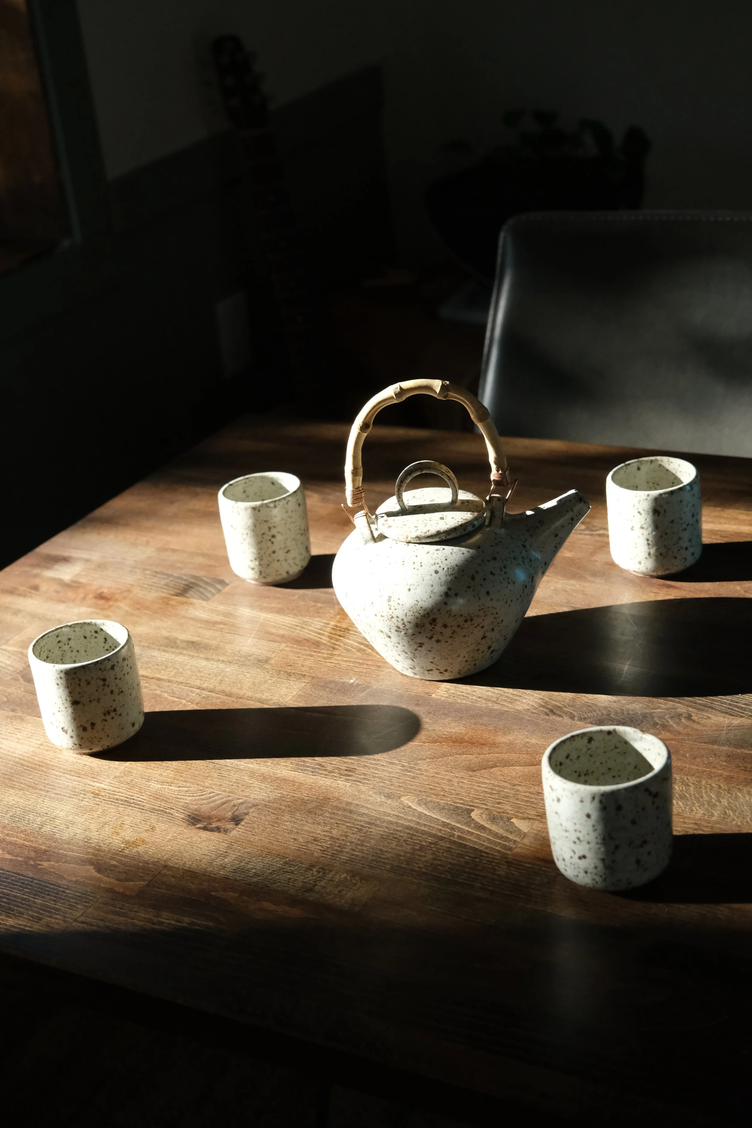 A white ceramic teapot with a bamboo handle and matching cups on a wooden table, with sunlight casting shadows.