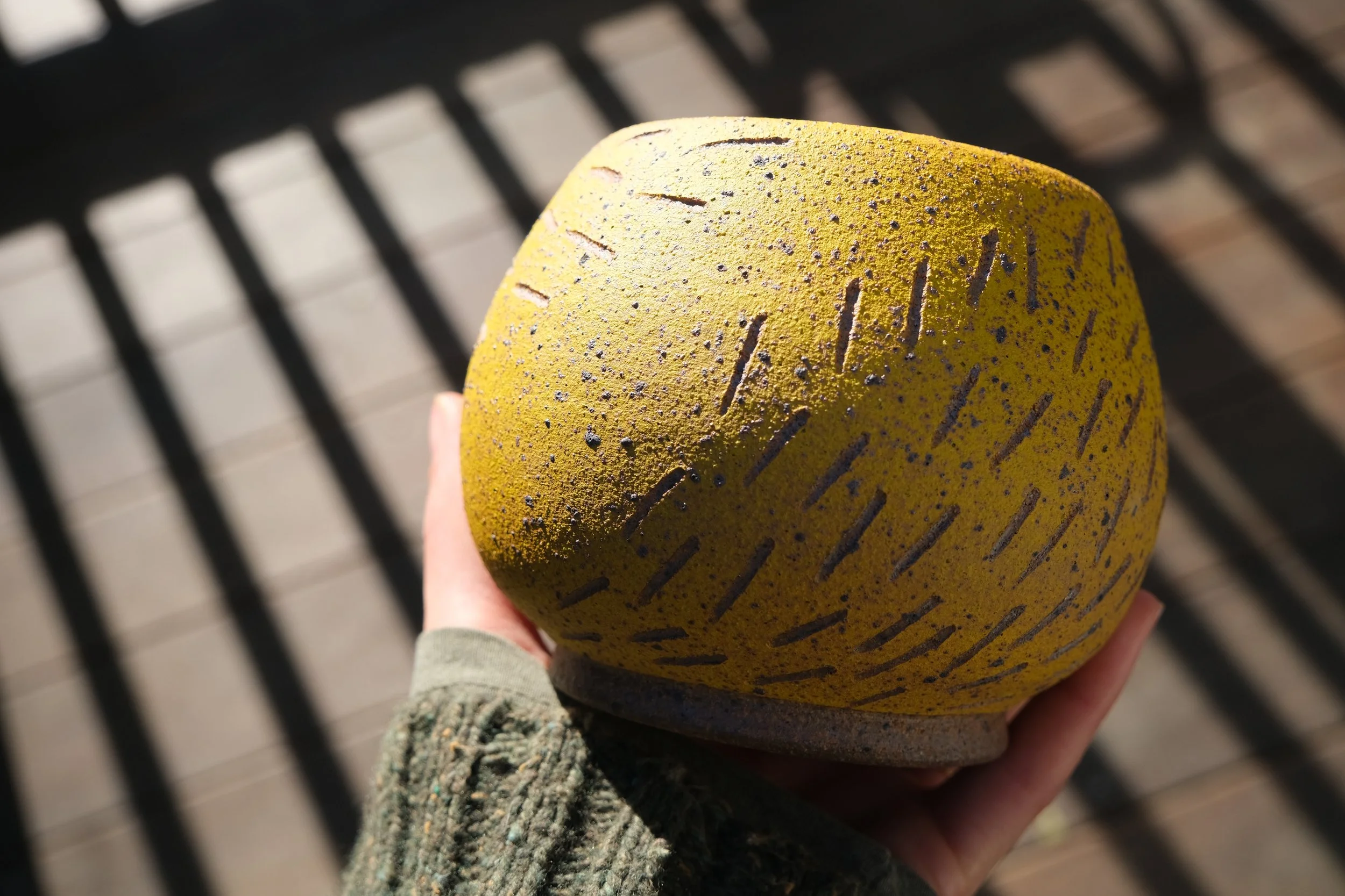 Close-up of a yellow climbing hold with black markings being held by a person wearing a greenish sweater, with a shadow-patterned metal surface in the background.