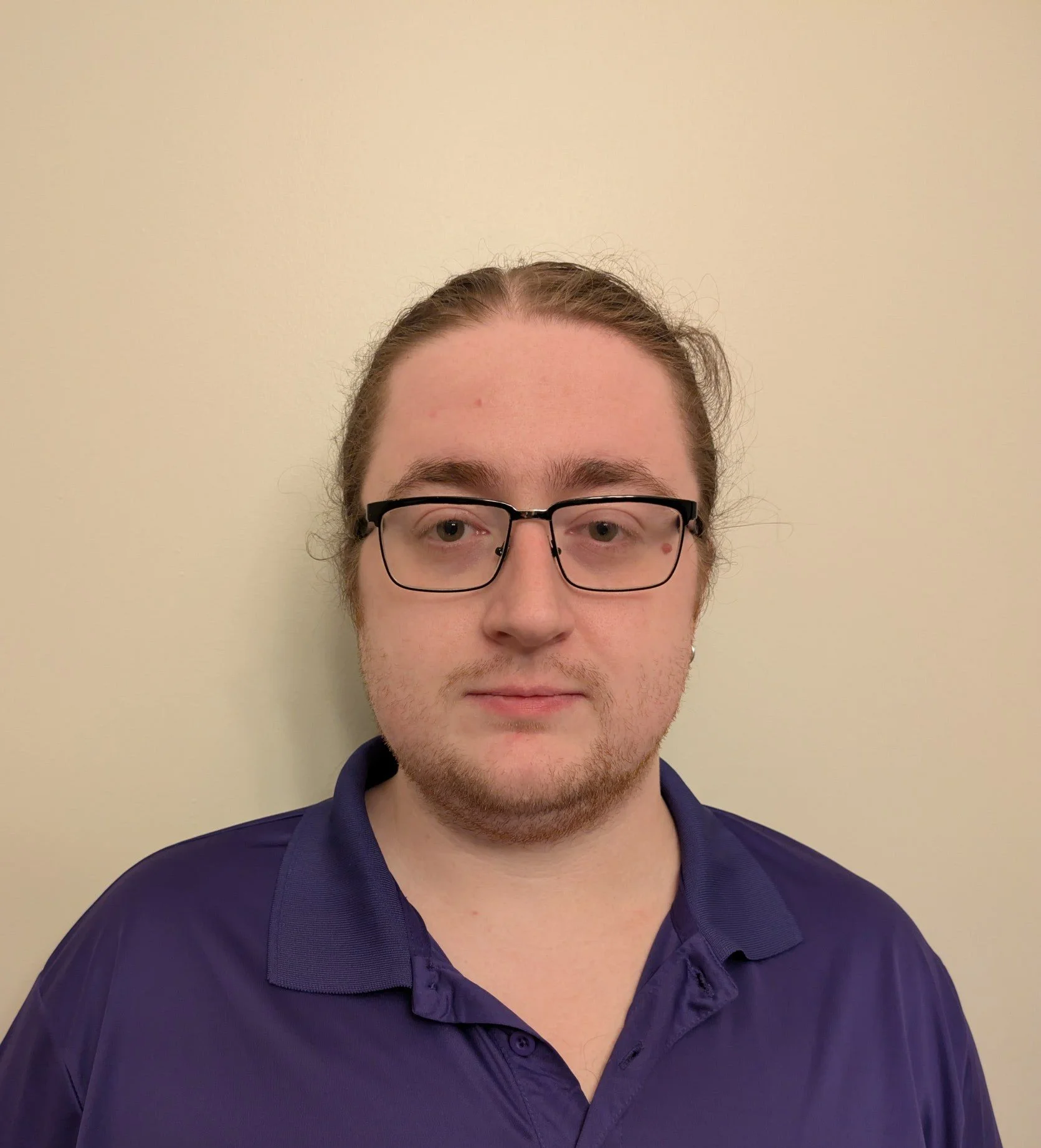 Close-up of a young man with short brown hair, glasses, and a beard, wearing a dark purple collared shirt, standing against a plain light-colored wall.