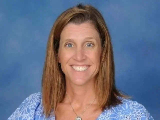 A woman with shoulder-length, reddish-brown hair, smiling, wearing a blue patterned top and a silver necklace, in front of a plain blue background.
