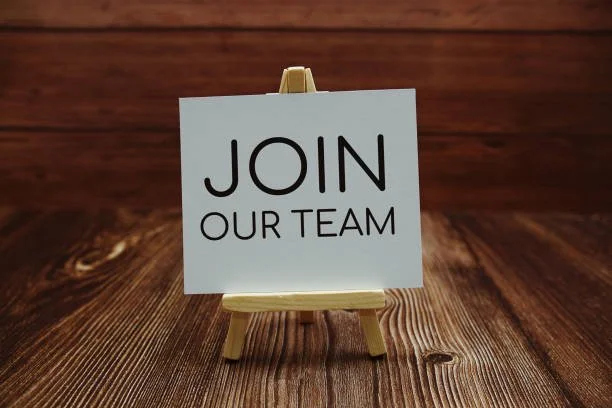 A small sign on a wooden stand displays the message 'Join Our Team' in black letters against a white background, on a polished wooden surface with a wooden wall in the background.