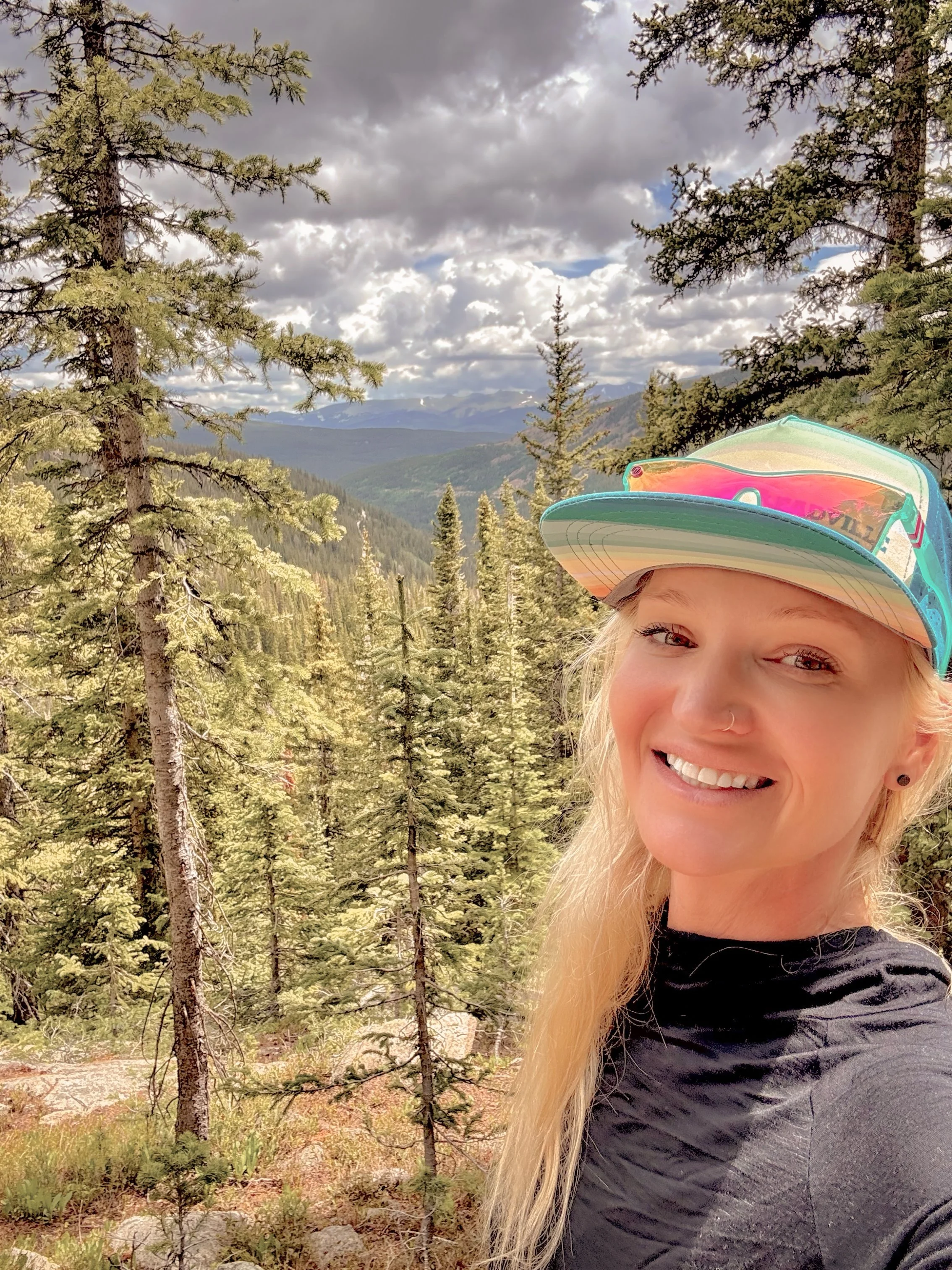 A young woman with blonde hair smiling outdoors among tall evergreen trees, wearing a colorful cap and a black shirt, with mountains and cloudy sky in the background.