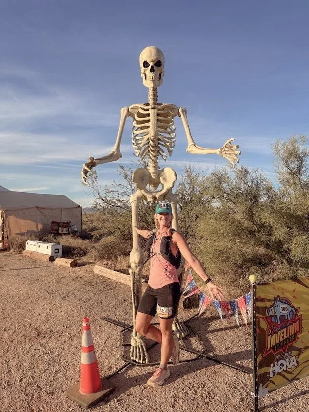 A woman standing next to a large skeleton statue outdoors in a desert setting, with a small flag and traffic cone nearby.