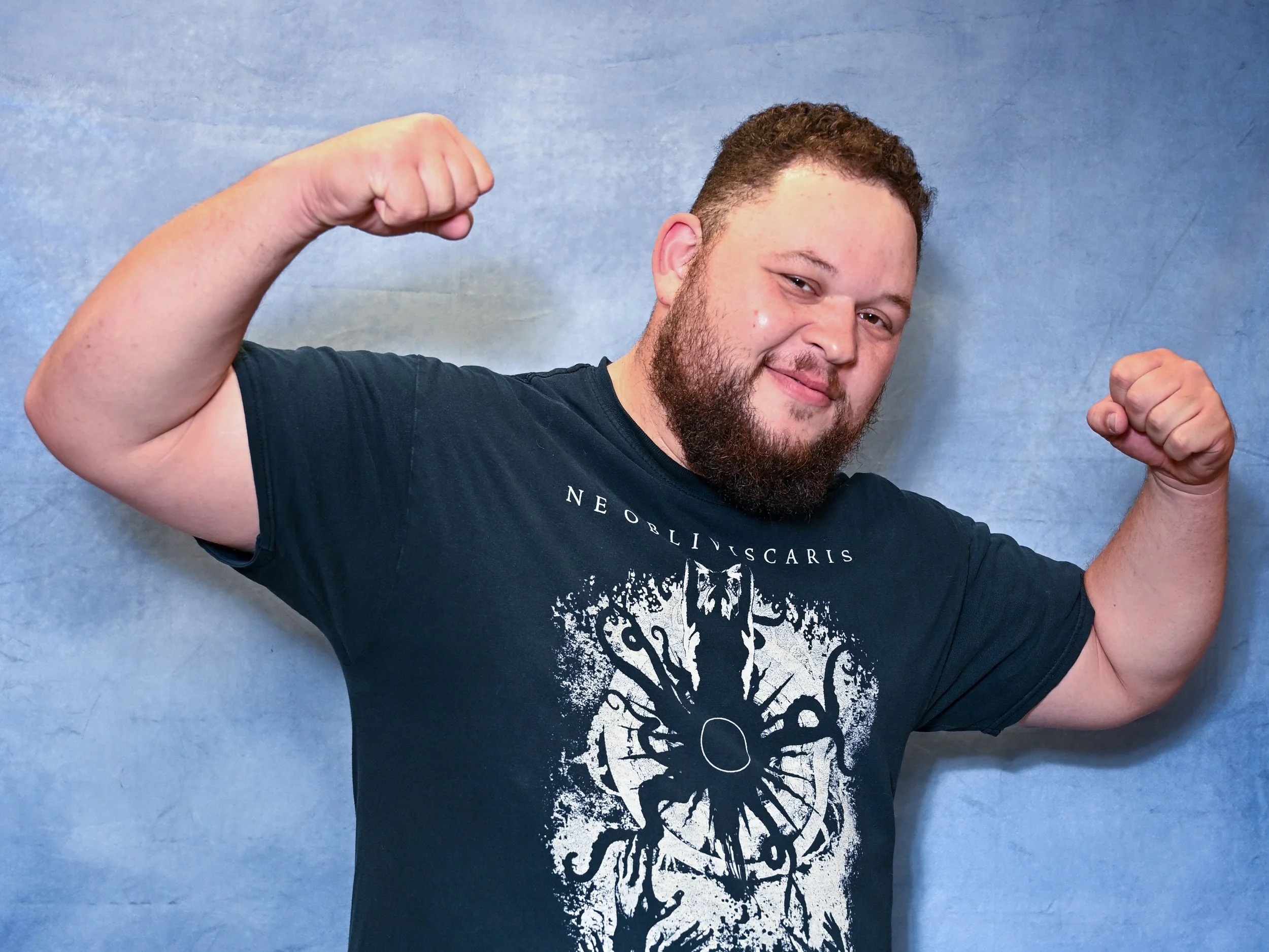 A man with a beard and short hair flexing his arm muscles, wearing a black T-shirt with a white graphic design, standing against a blue textured background.