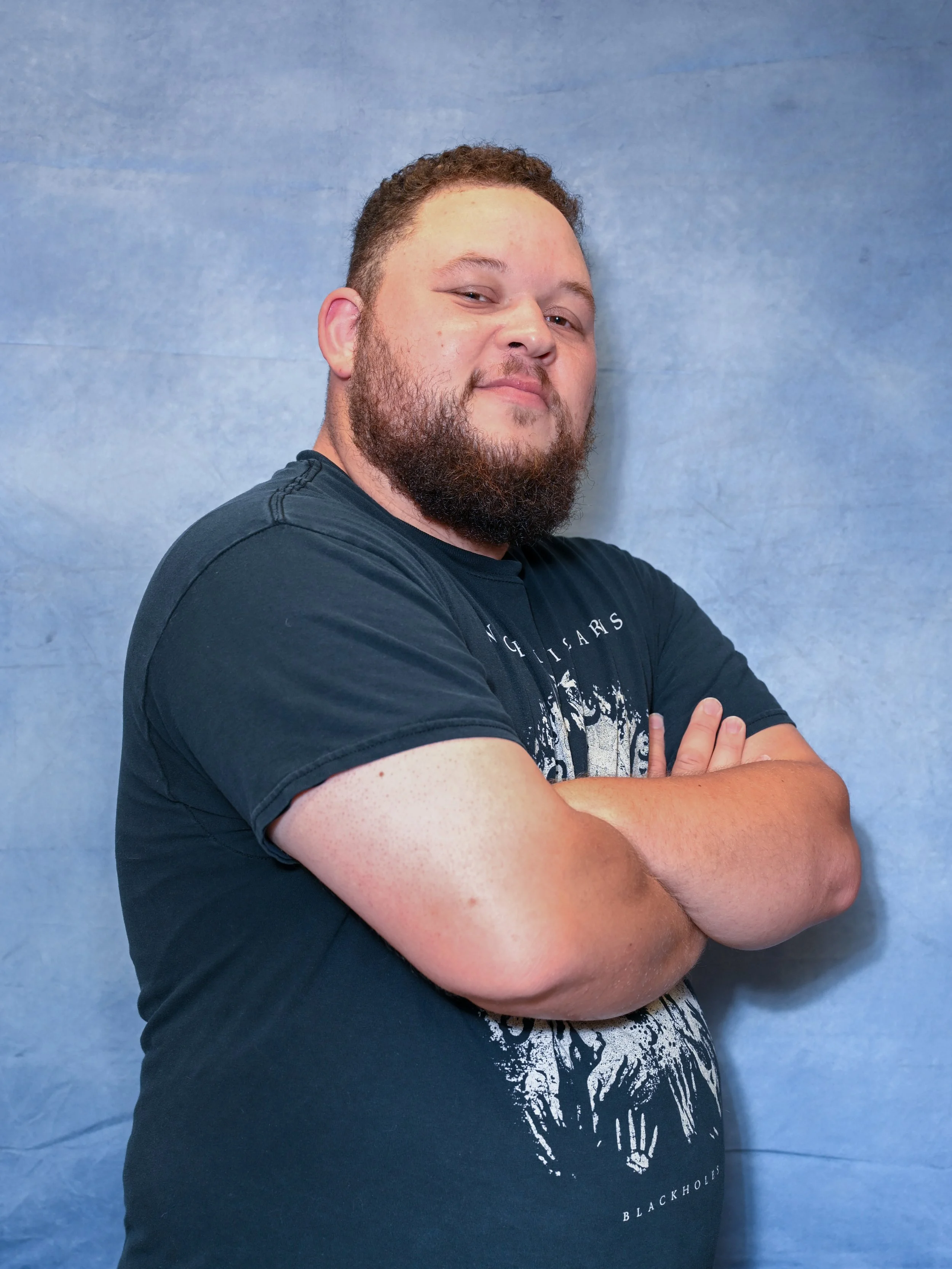 A man with a beard and short curly hair standing with arms crossed in front of a blue background. He is wearing a black t-shirt with white graphic text and design.