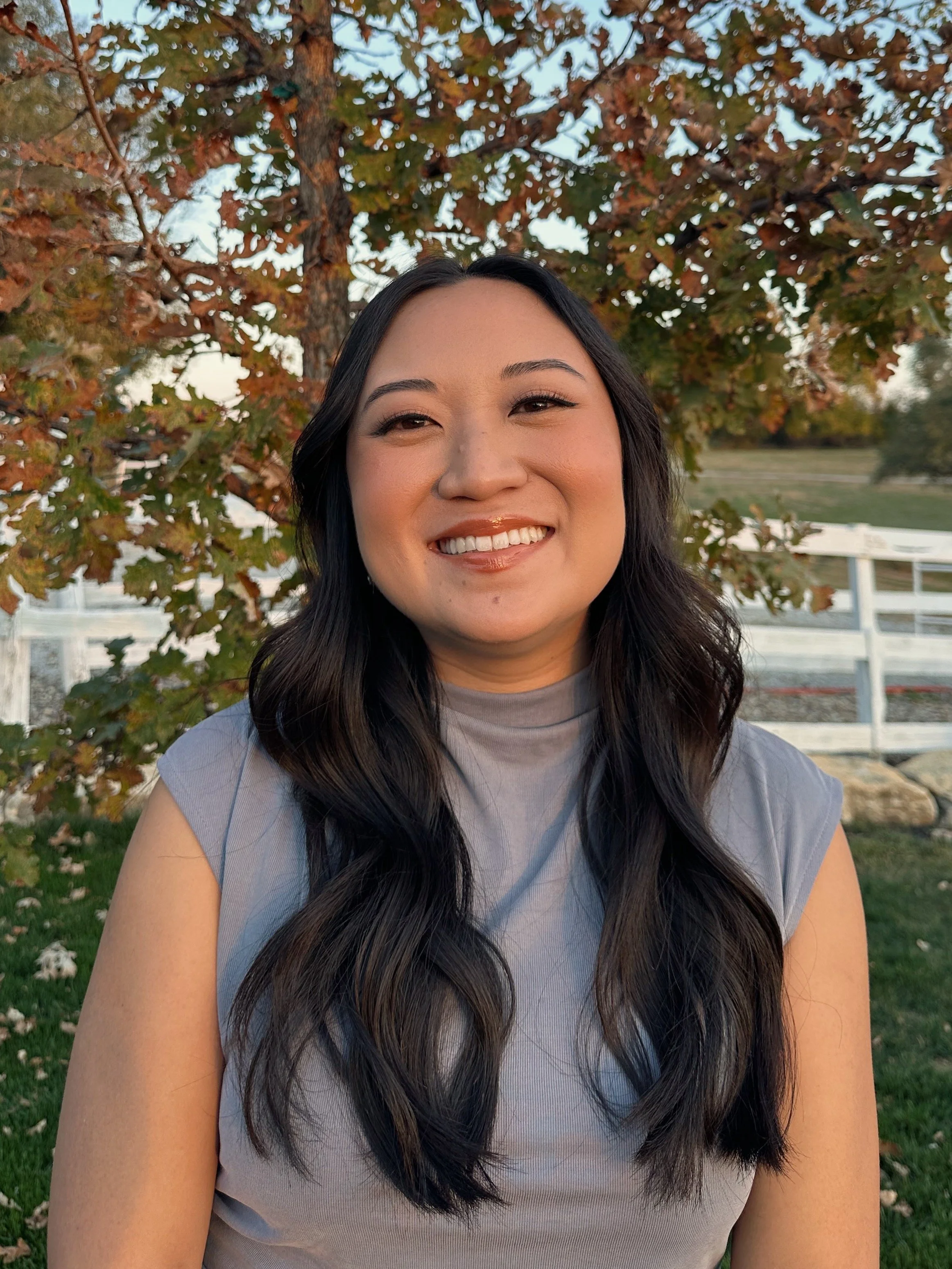 Close-up of a smiling woman with long dark hair, standing outdoors in front of a tree with autumn-colored leaves, a white fence, and a grassy area in the background.