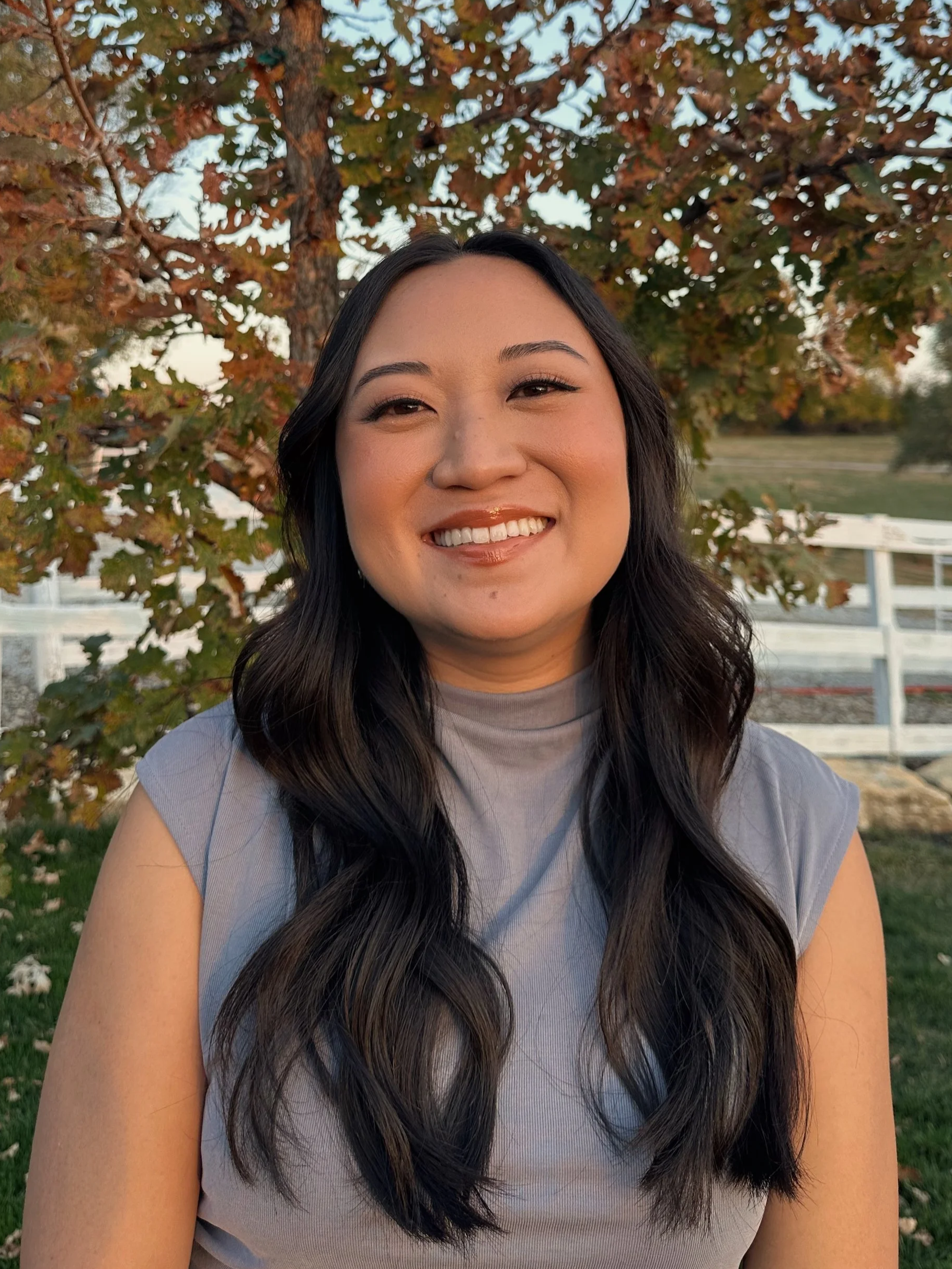 A woman with long dark hair smiling outdoors with a tree and a white fence in the background.